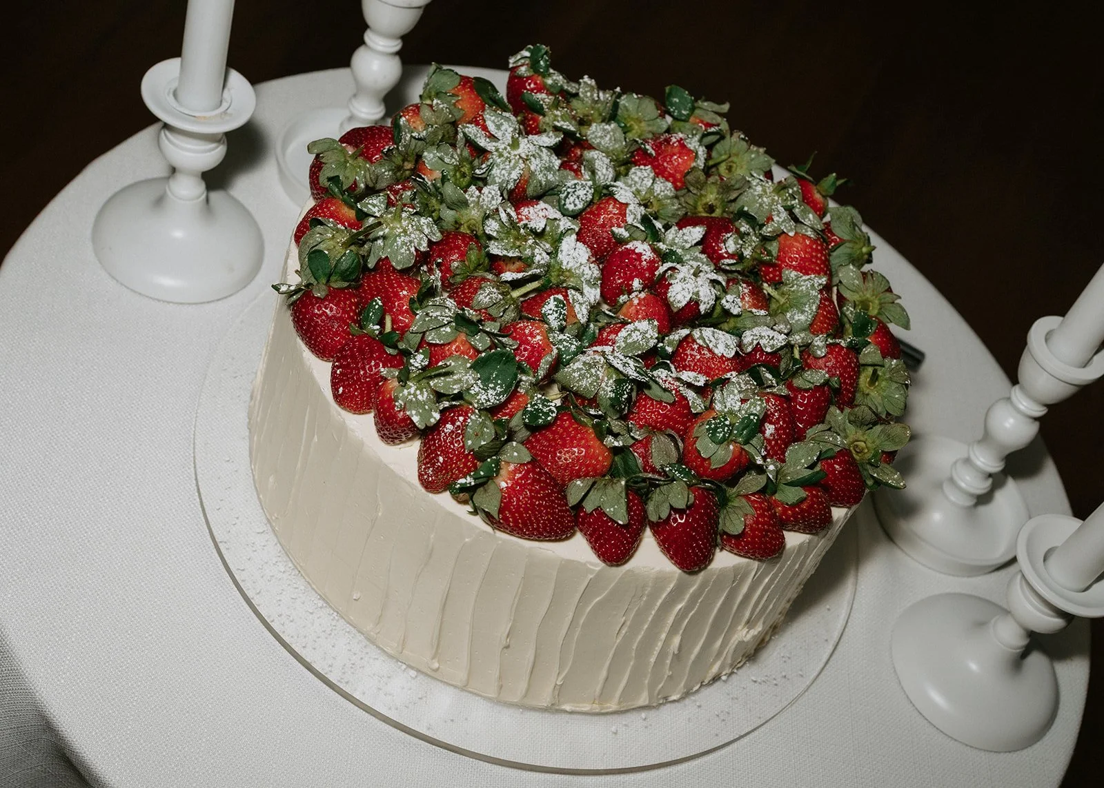 A round cake covered with white frosting, topped with fresh strawberries, green leaves, and a light dusting of powdered sugar, placed on a white tablecloth with white candlesticks around it.