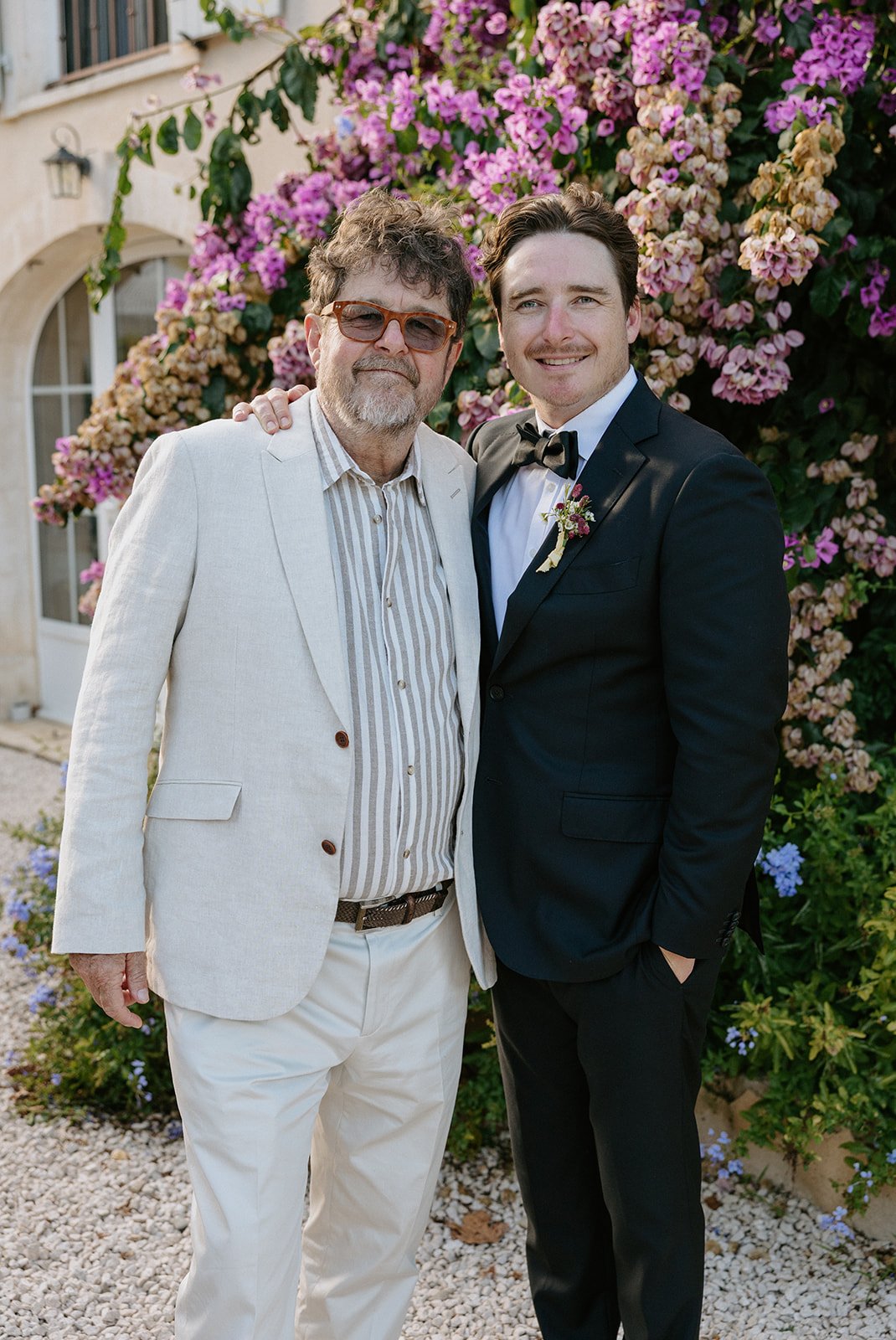 A man in a light-colored suit and a man in a black tuxedo pose together outdoors in front of blooming pink and purple flowers.