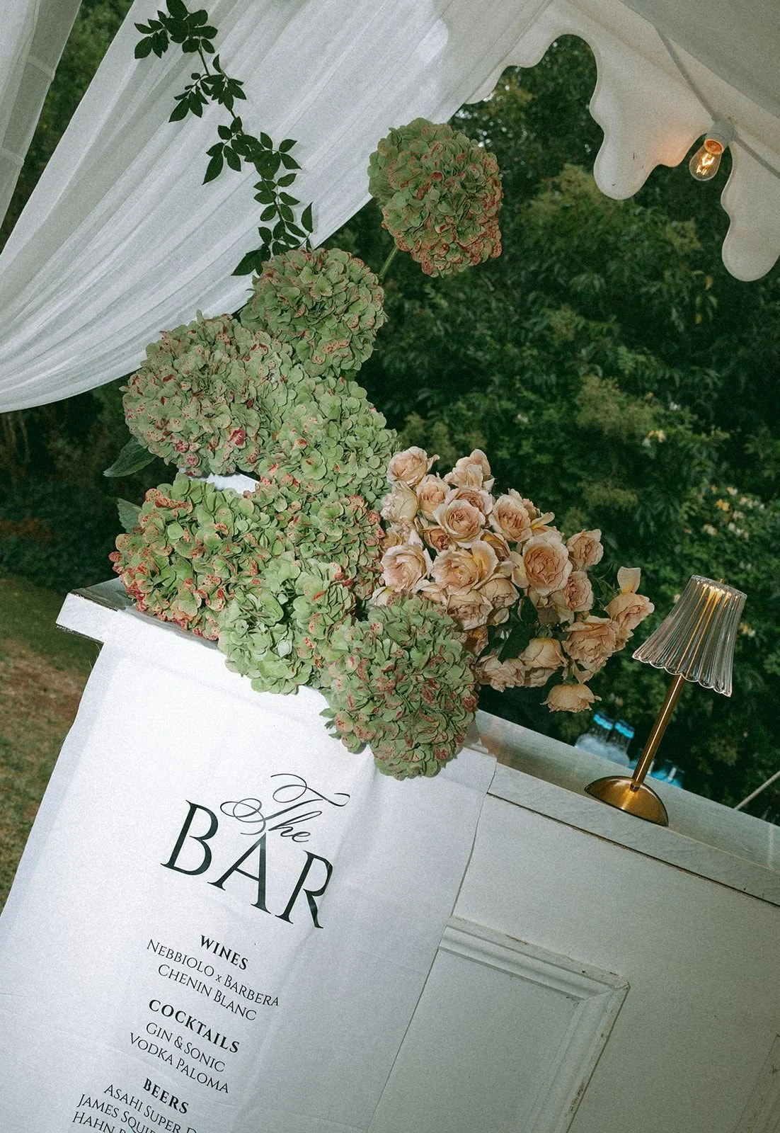 A white bar sign with black text listing wines, cocktails, and beers, next to a floral arrangement of pink roses and green hydrangeas, with a small gold lamp beside it, under a white canopy with sheer curtains at an outdoor event.