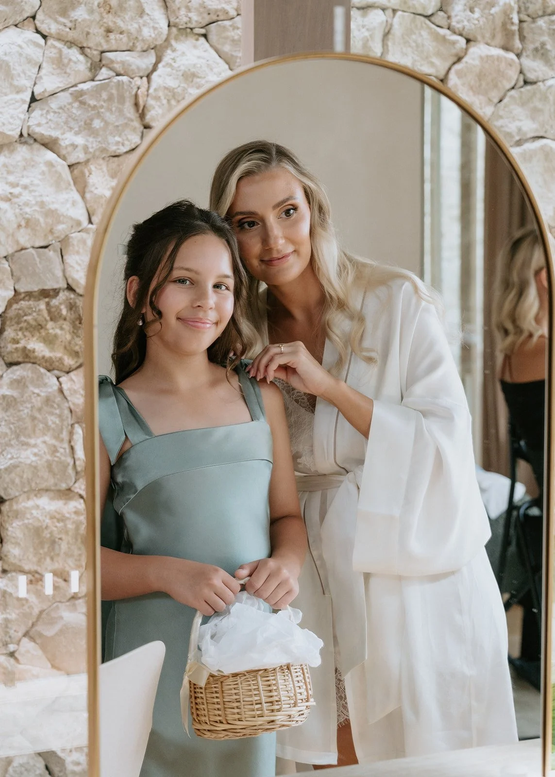 A woman and a girl pose for a photo in a mirror, with a stone wall background. The girl is holding a wicker basket with tissue paper, and the woman is touching her shoulder.