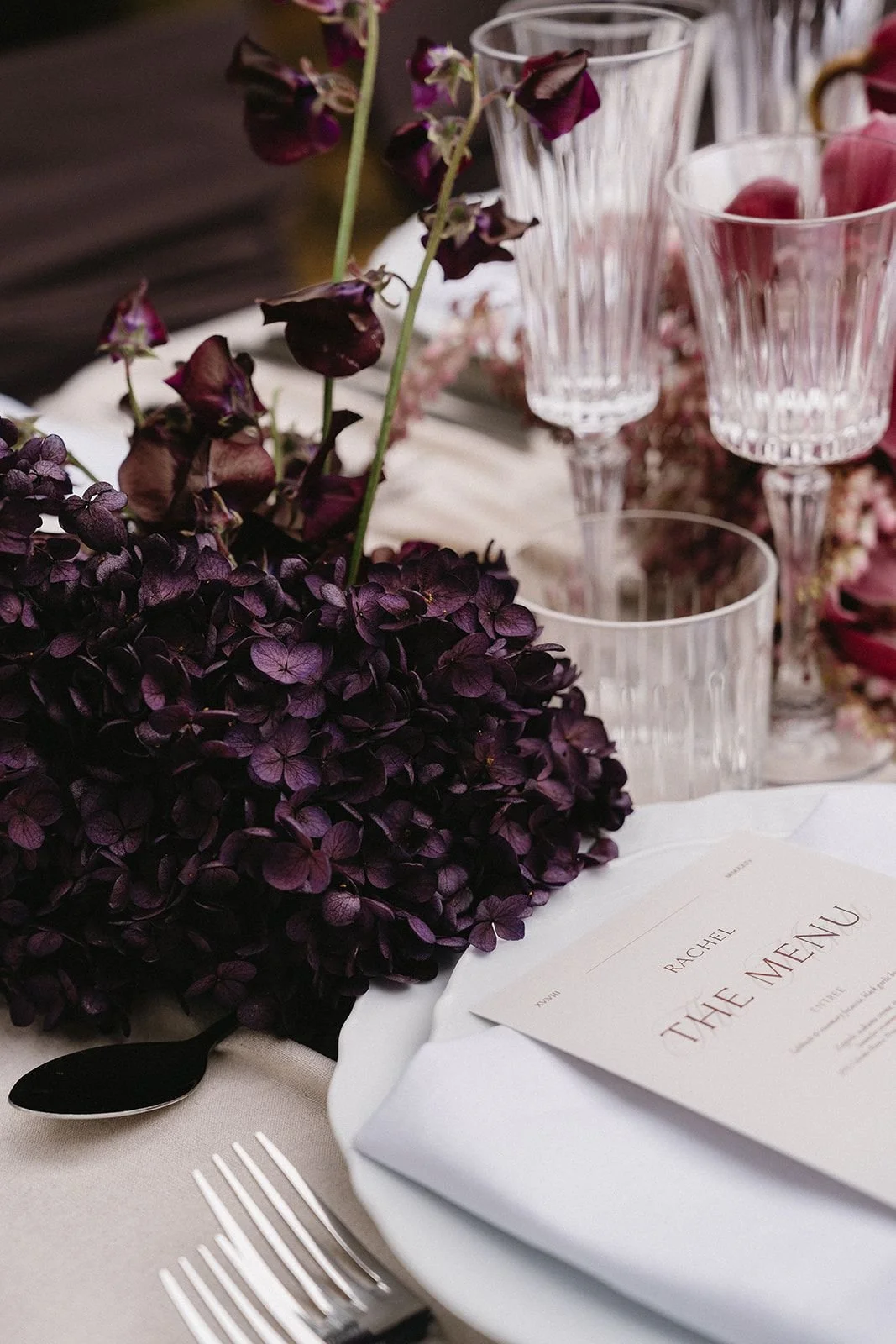 Elegant table setting with dark purple hydrangeas, clear glassware, a white plate with a folded napkin, silverware, and a menu titled 'Rachel The Menu' on a beige tablecloth.