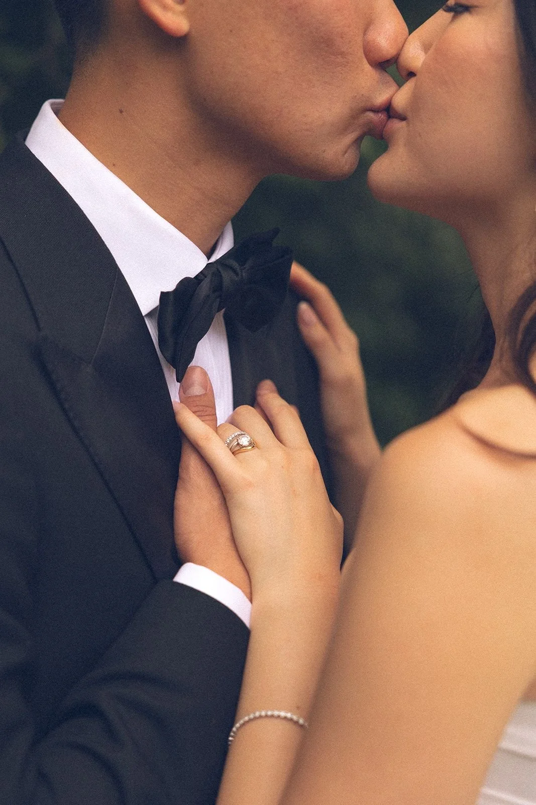 A close-up of a couple kissing, dressed in formal wedding attire, with the woman's hand resting on the man's chest, showing a wedding ring and a bracelet.