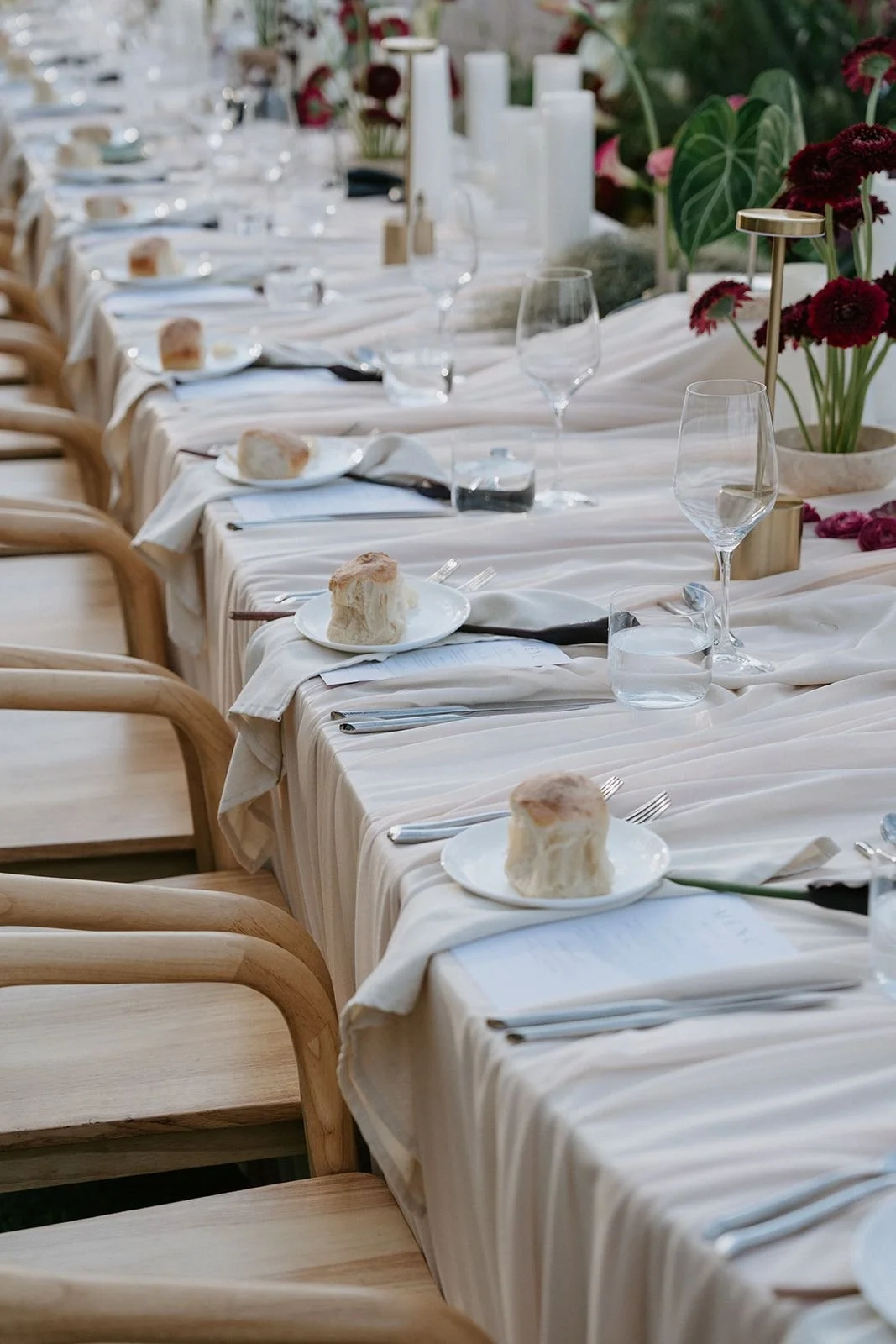Long dining table set with white tablecloths, wine glasses, plates of bread, and cutlery, decorated with large red flowers and greenery.