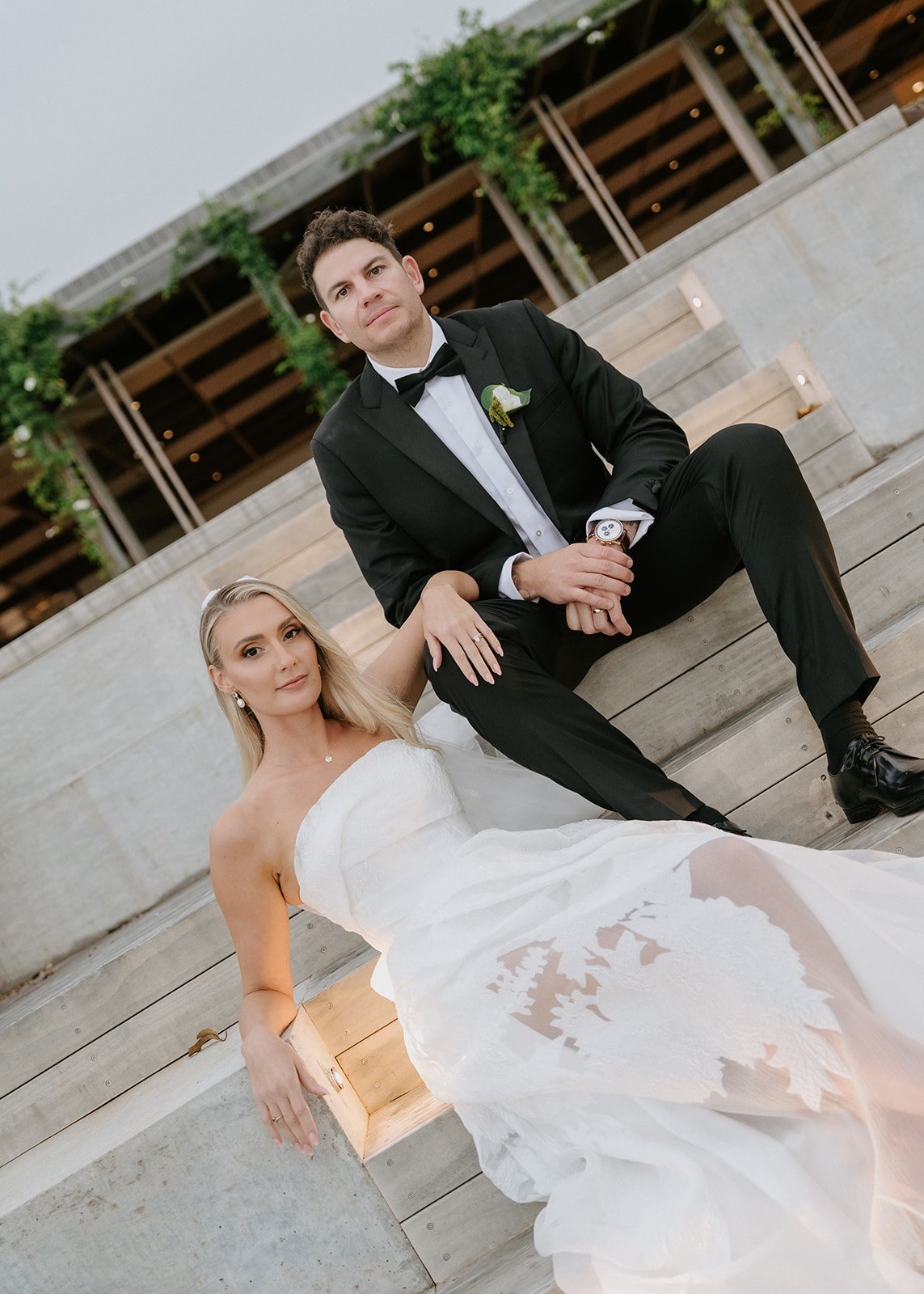 A newlywed couple, the bride in a strapless white wedding gown and the groom in a black tuxedo, sitting on wooden steps outdoors during daytime. The bride has long blonde hair and is wearing earrings, while the groom has dark hair and is wearing a wr