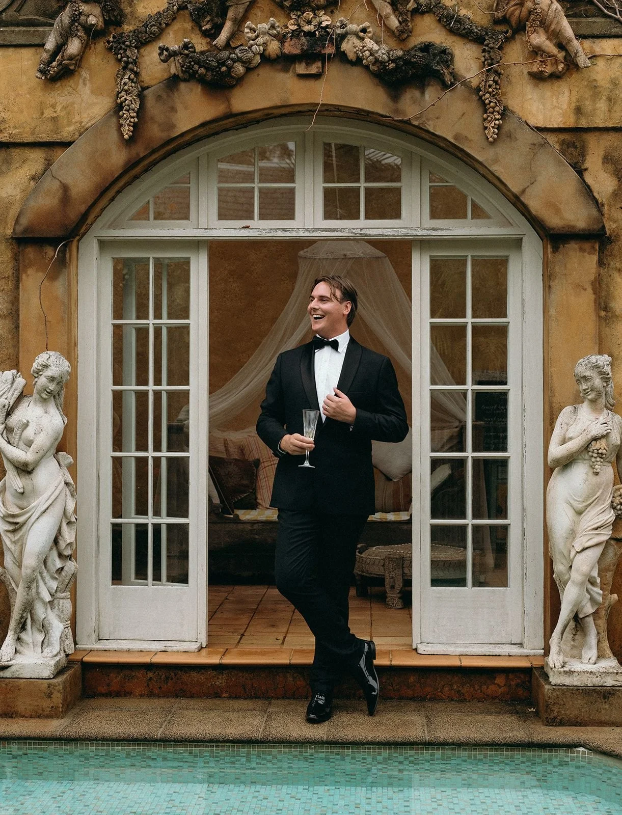 A man in a tuxedo holding a glass of champagne, standing by a doorway with glass panes, smiling, with a wedding veil in the background.