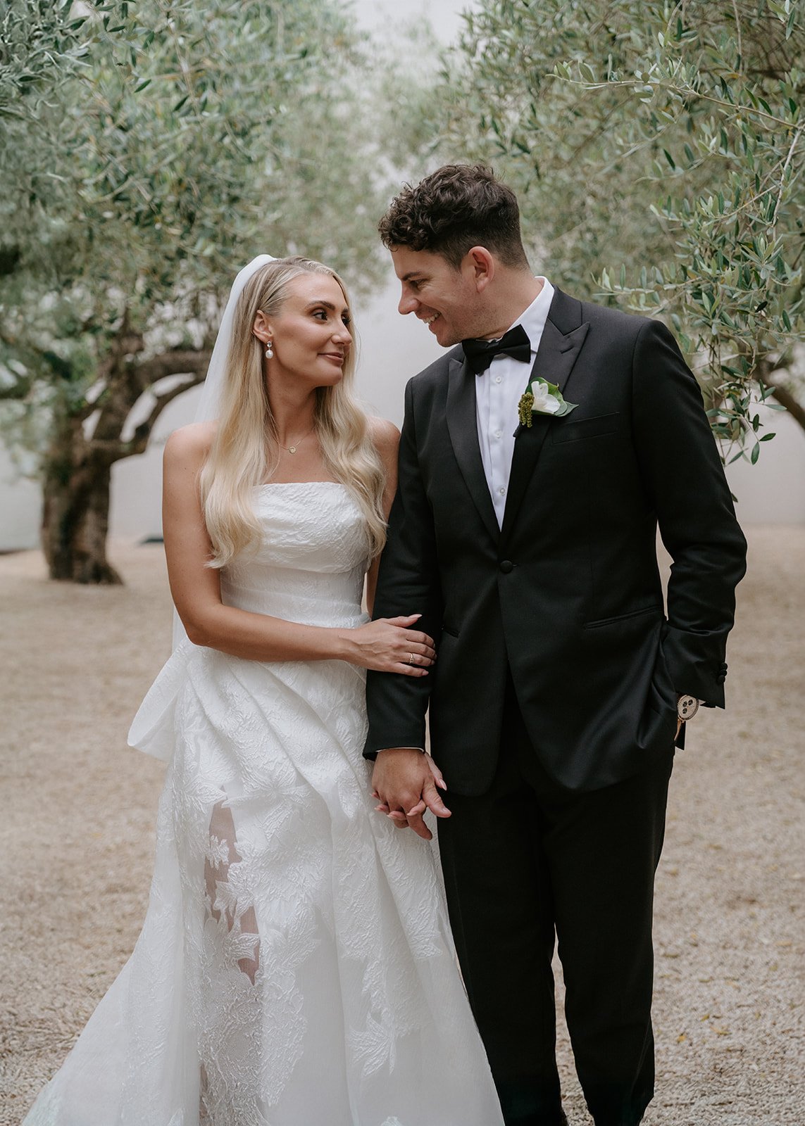 A bride and groom hold hands, standing outdoors under trees, gazing into each other's eyes. The bride wears a strapless white wedding gown with lace details and a veil, and the groom wears a black tuxedo with a bow tie and boutonnière.
