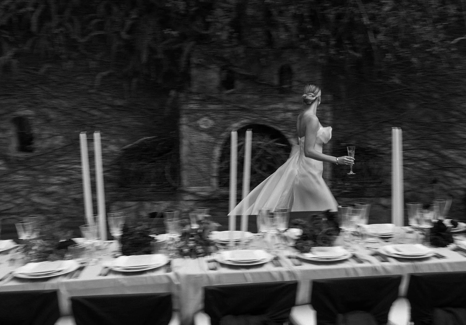 A woman in a strapless wedding dress walking along a table set for a celebration, holding a glass of champagne in her right hand, with dark outdoor scenery in the background.