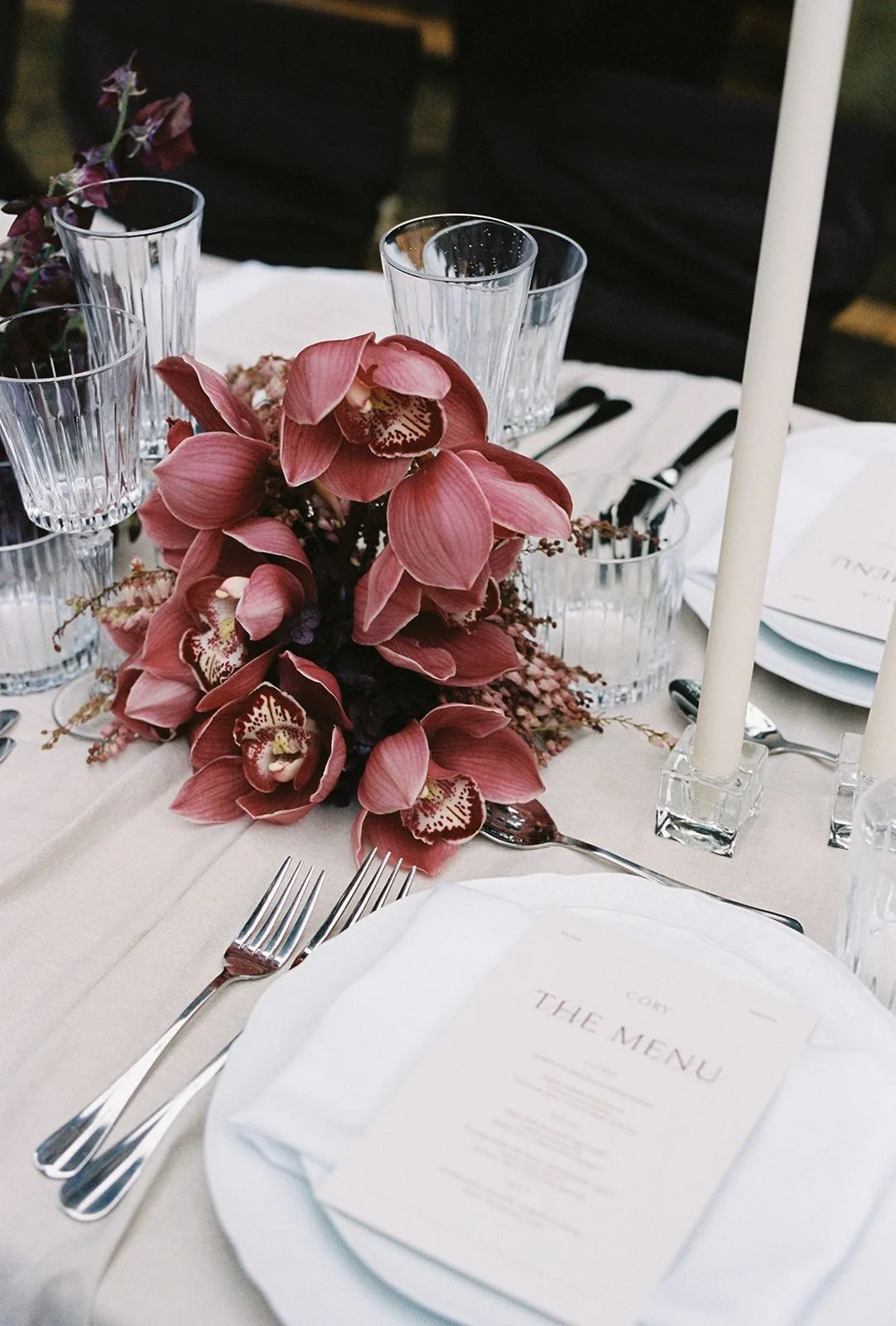 Elegant table setting with pink orchid floral centerpiece, white napkins, glassware, silverware, and a menu card, featuring two candles in glass holders.