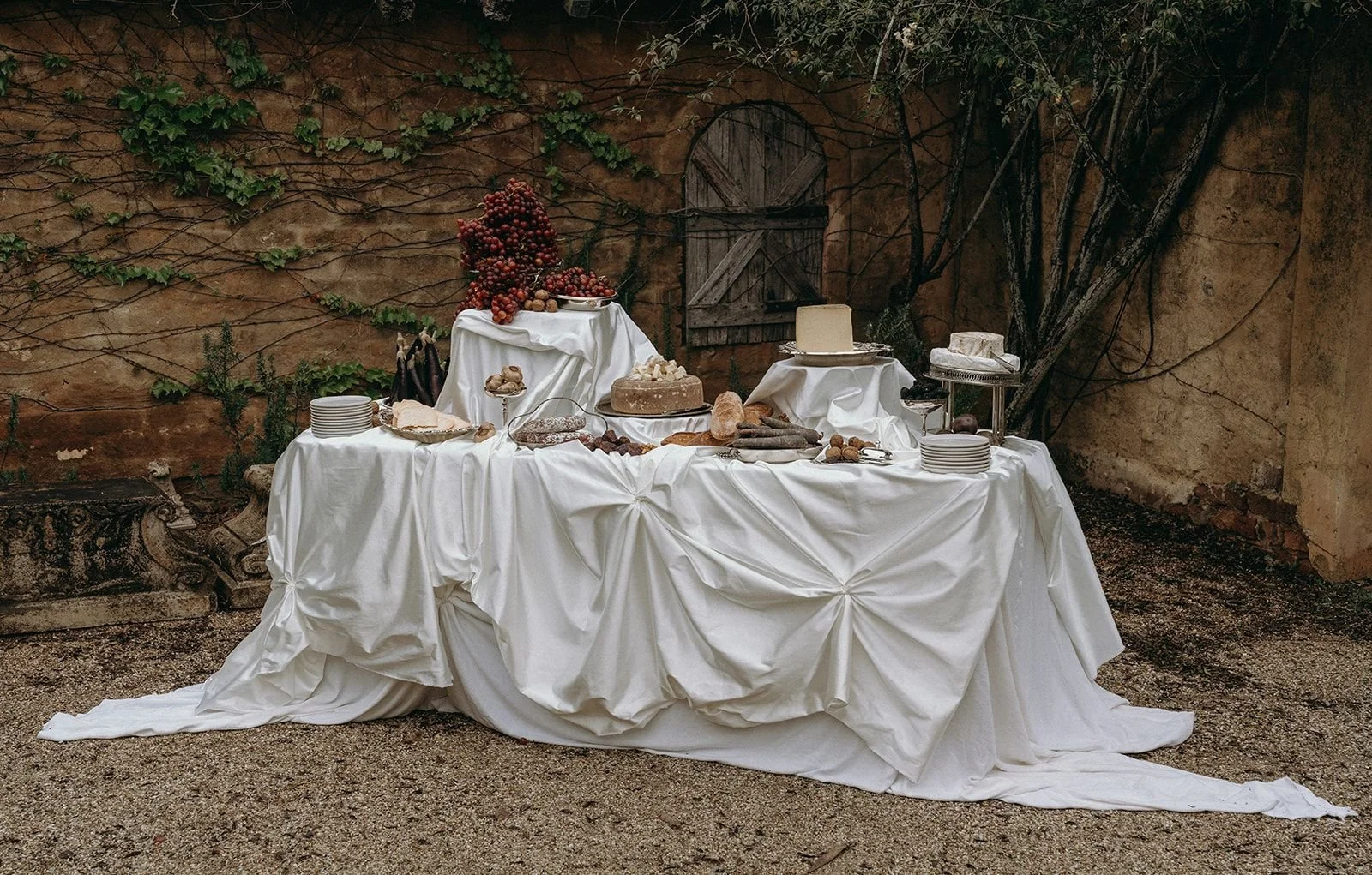 A rustic outdoor dessert table covered with white fabric, set against a weathered brick wall with vines, holding grapes, cakes, and plates of food.
