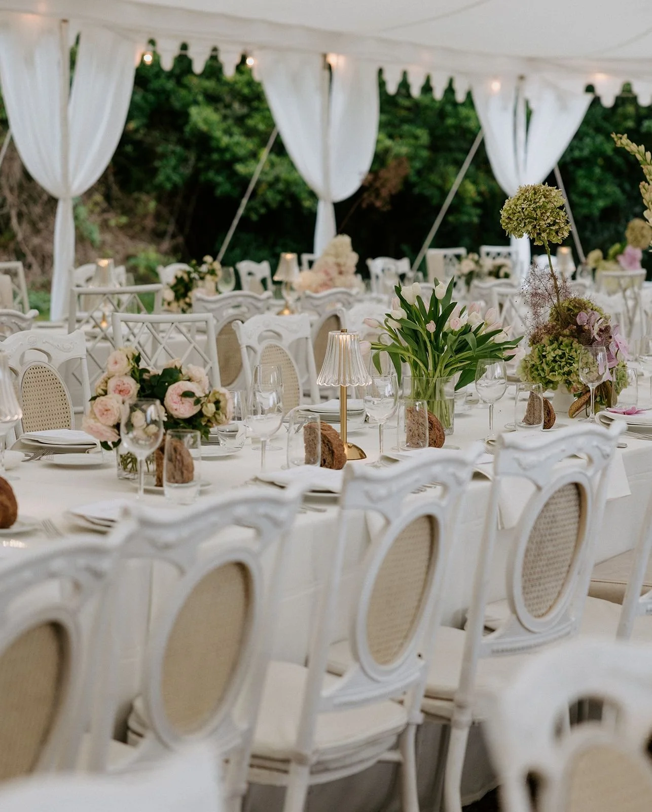 Elegant outdoor event setup with long white tables, white chairs, pink and green floral centerpieces, candles, and soft lighting in a white tent with curtains, greenery in the background.