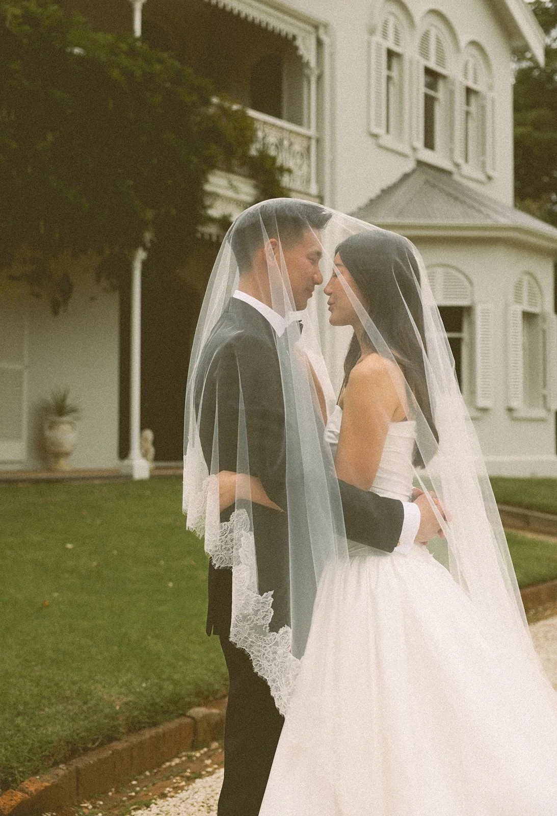 Bride and groom standing close under a veil in front of a Victorian-style house