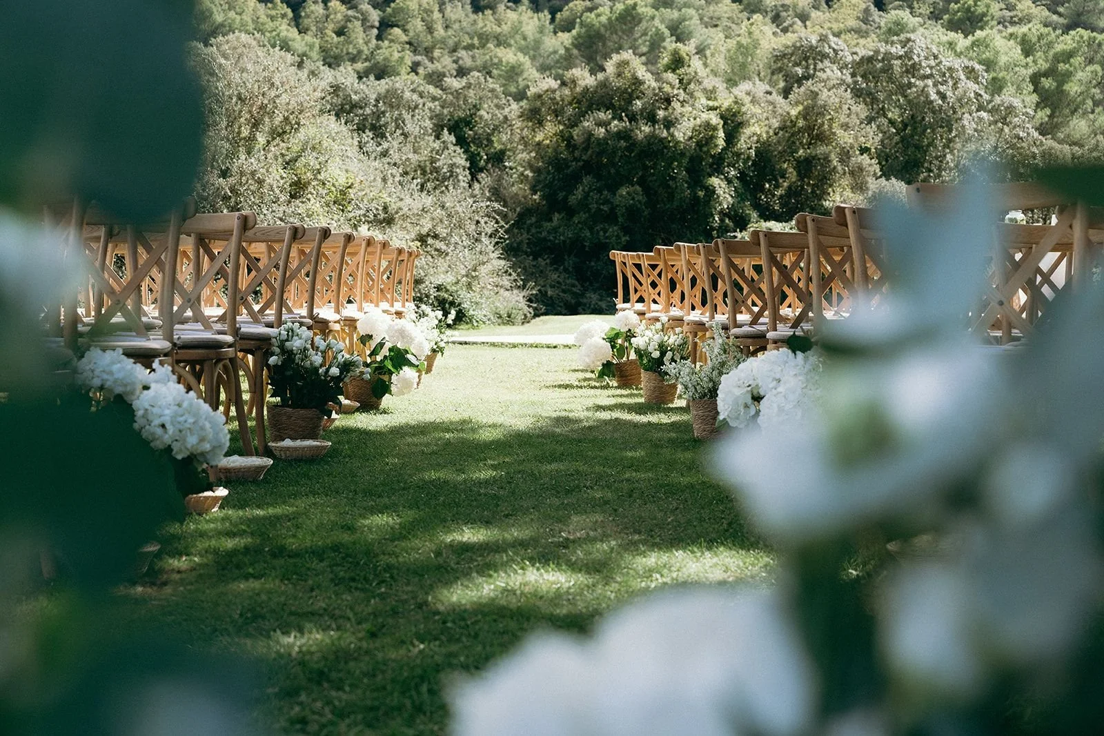 Outdoor wedding setup with wooden chairs and white floral arrangements along a grassy aisle, surrounded by trees.