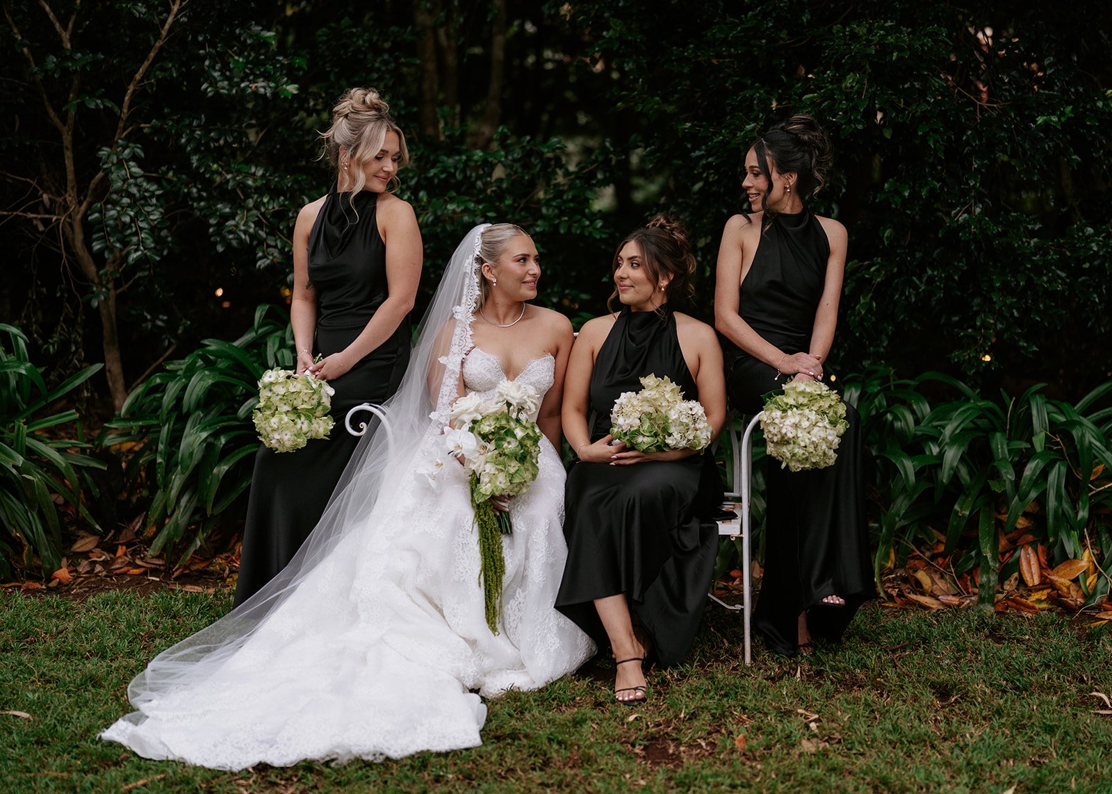 A bride in a white wedding gown with a lace veil and three bridesmaids in black dresses, holding bouquets of white flowers, sitting outdoors on a white bench in a garden with green plants and trees.