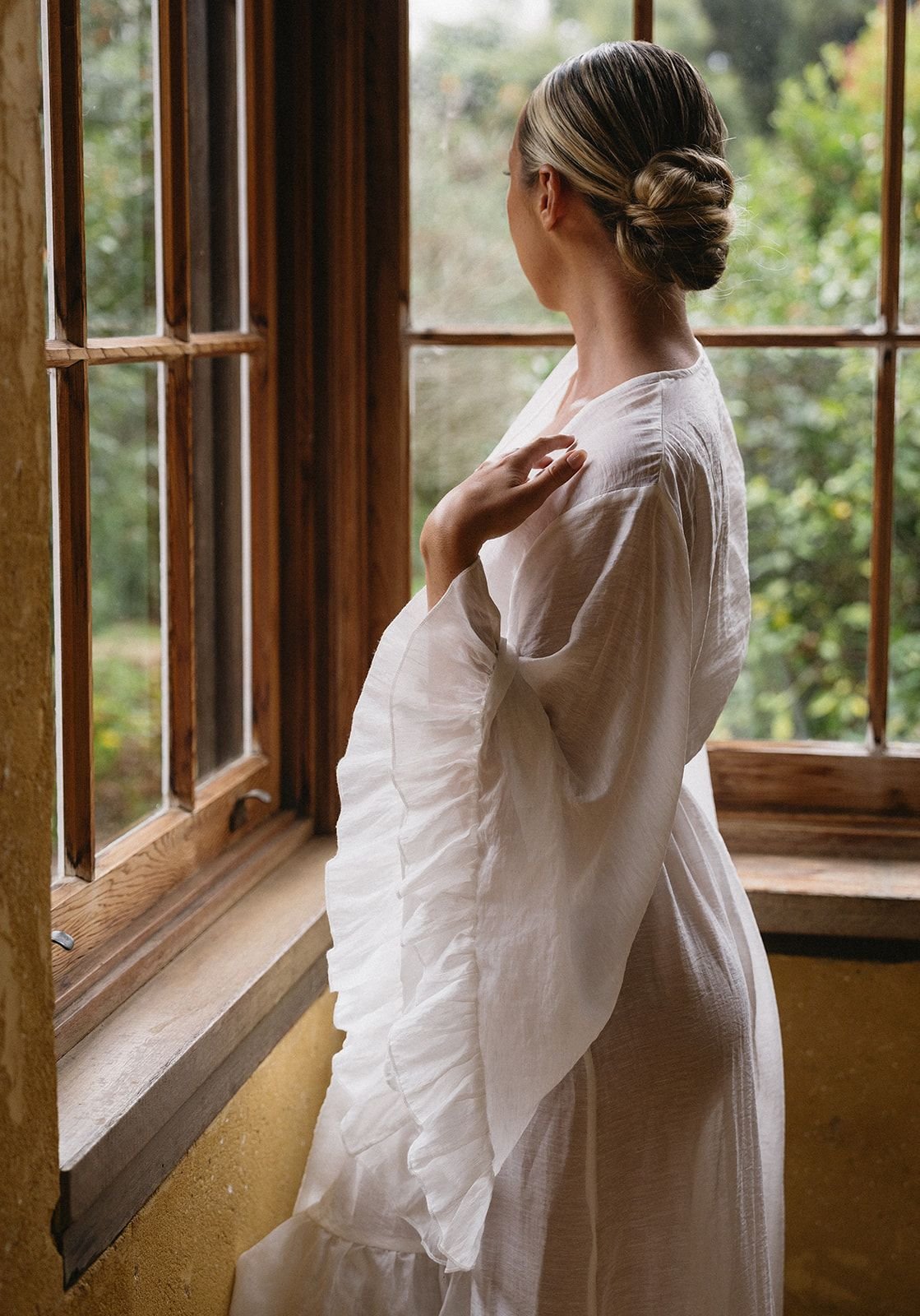 A woman with braided hair wearing a white dress, looking out a wooden-framed window onto greenery.