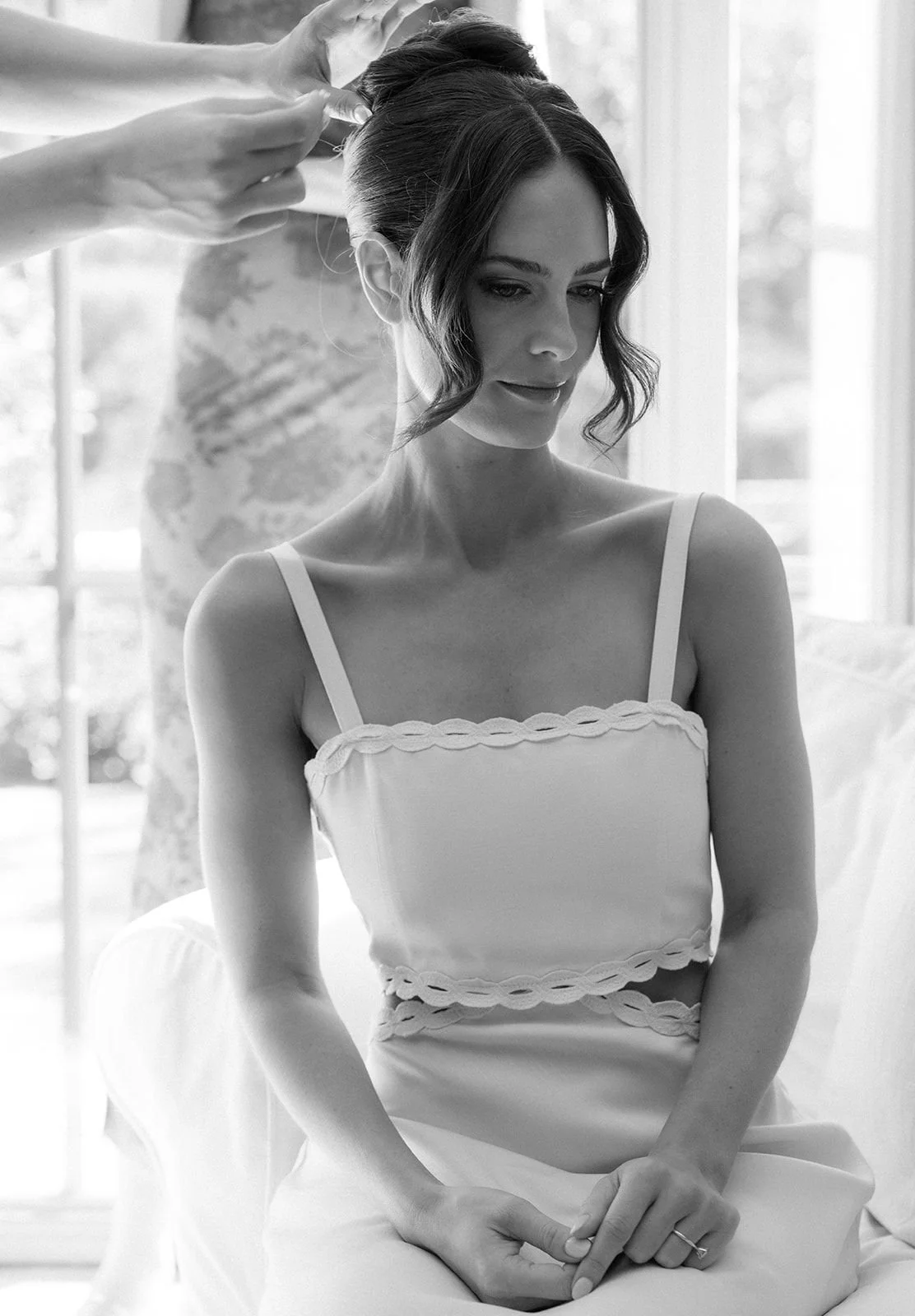 A woman has her hair styled by a hairstylist, sitting in front of a window in a bright room.