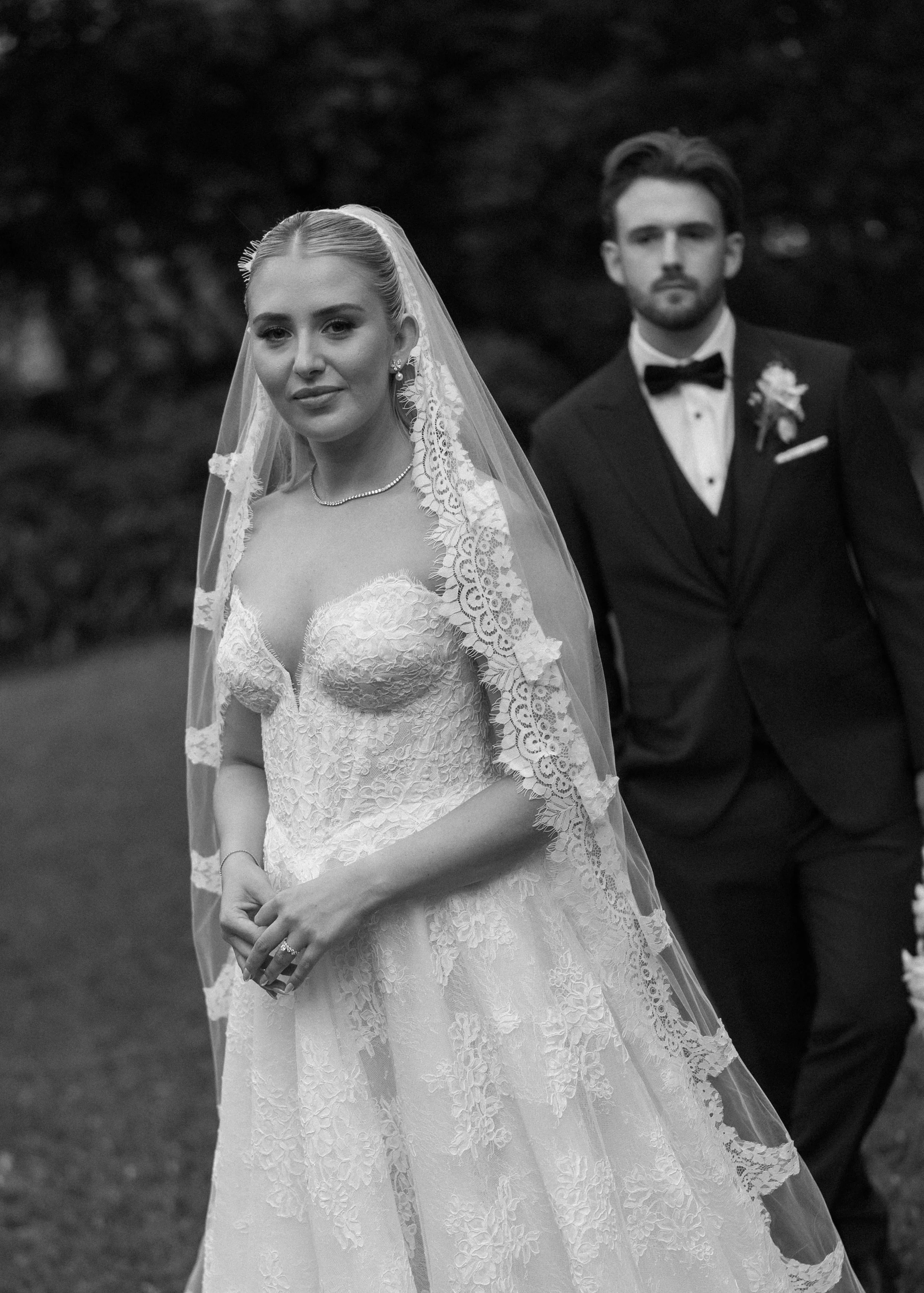 Black and white photograph of a bride in a lace wedding gown with a veil and a groom in a tuxedo with a bow tie, standing outdoors.