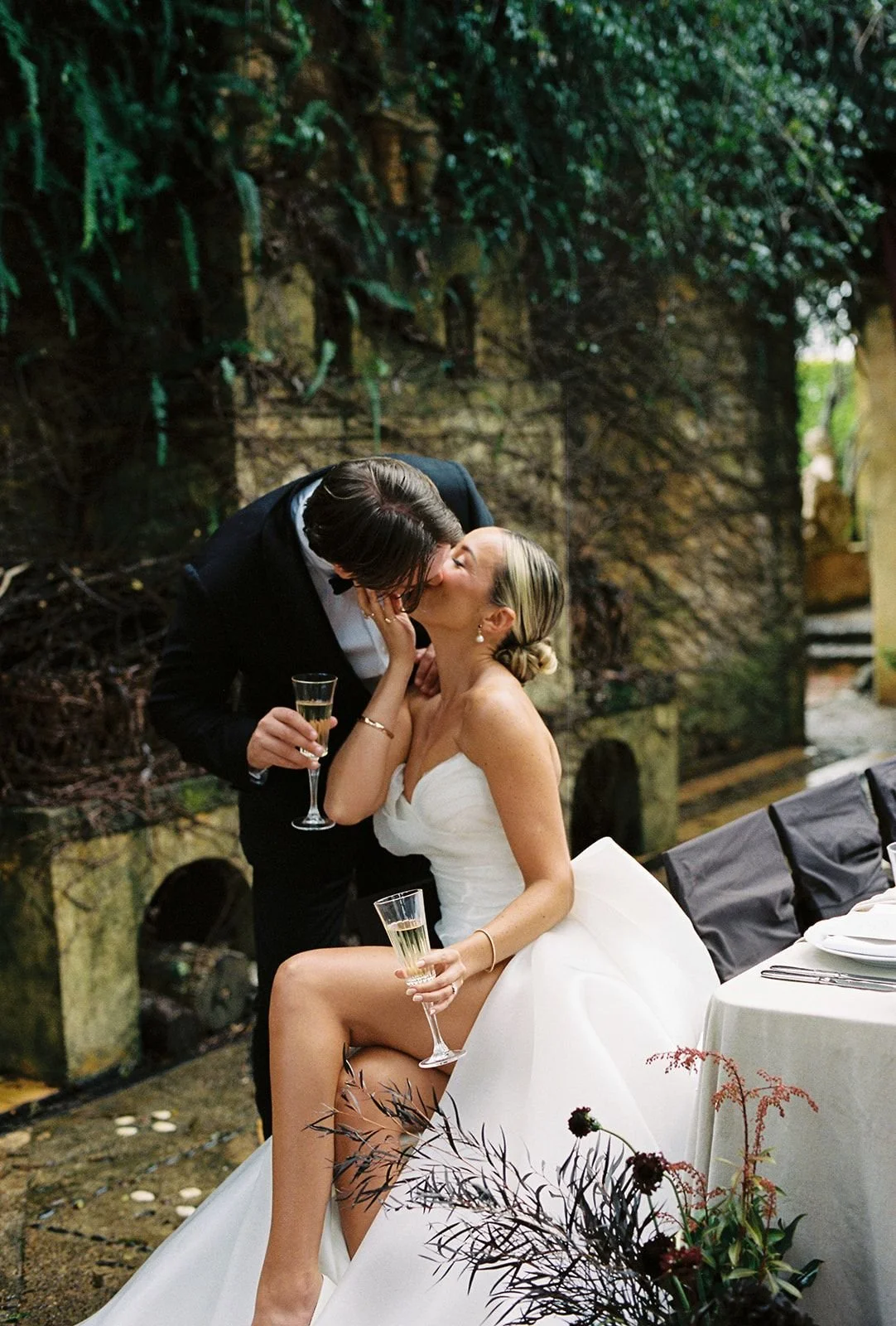 A newlywed couple sharing a kiss at their wedding reception, with the bride sitting on the table holding a glass of champagne, and the groom leaning in for the kiss, in an outdoor setting with greenery and a stone backdrop.