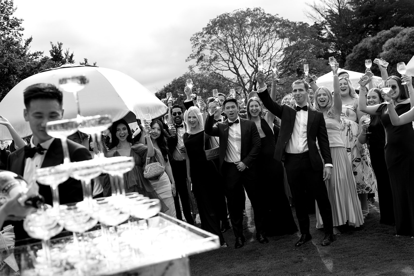 A group of wedding guests dressed in formal attire celebrating outdoors, holding glasses of champagne and smiling.