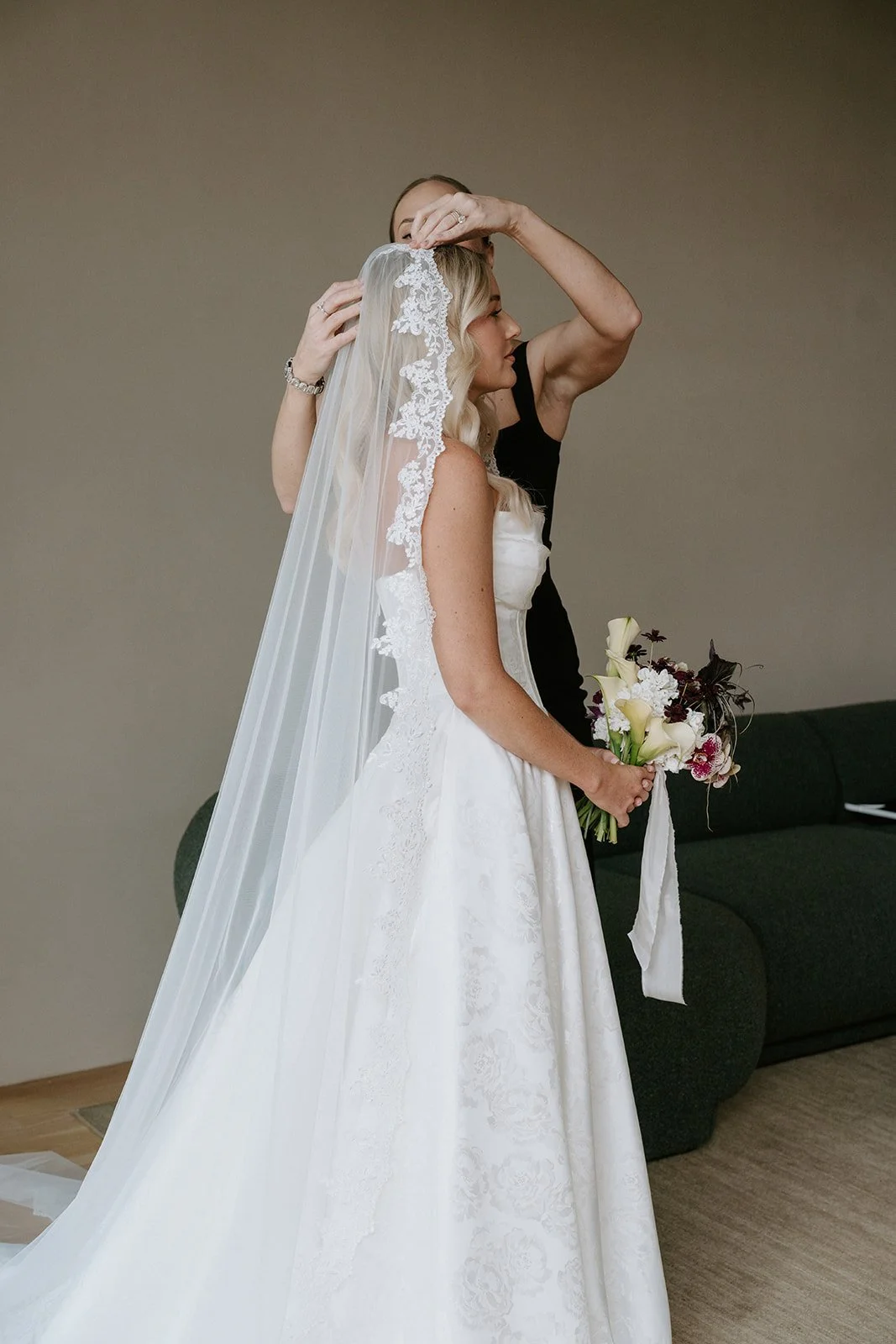 A bride with blonde hair wearing a white wedding dress and veil holding a bouquet of flowers, while another woman adjusts her veil.