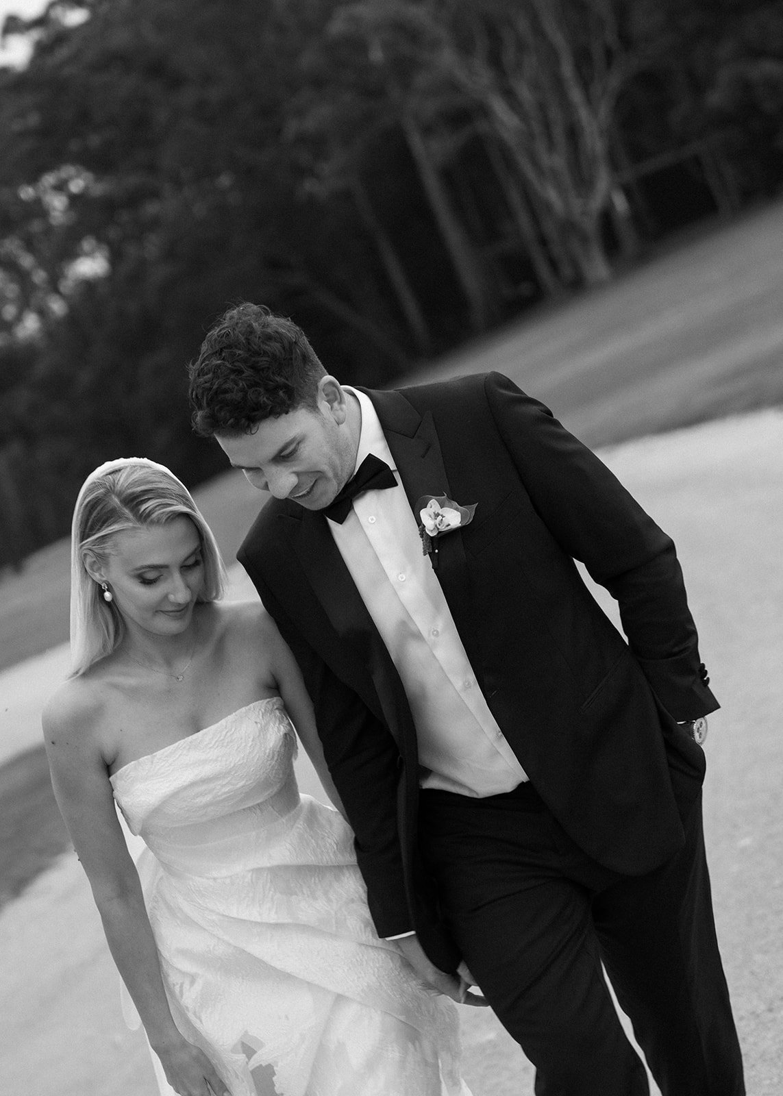Black and white photo of a bride and groom walking outdoors in a park, looking down, with trees in the background. The groom is wearing a tuxedo and bow tie, and the bride is wearing a strapless wedding dress.