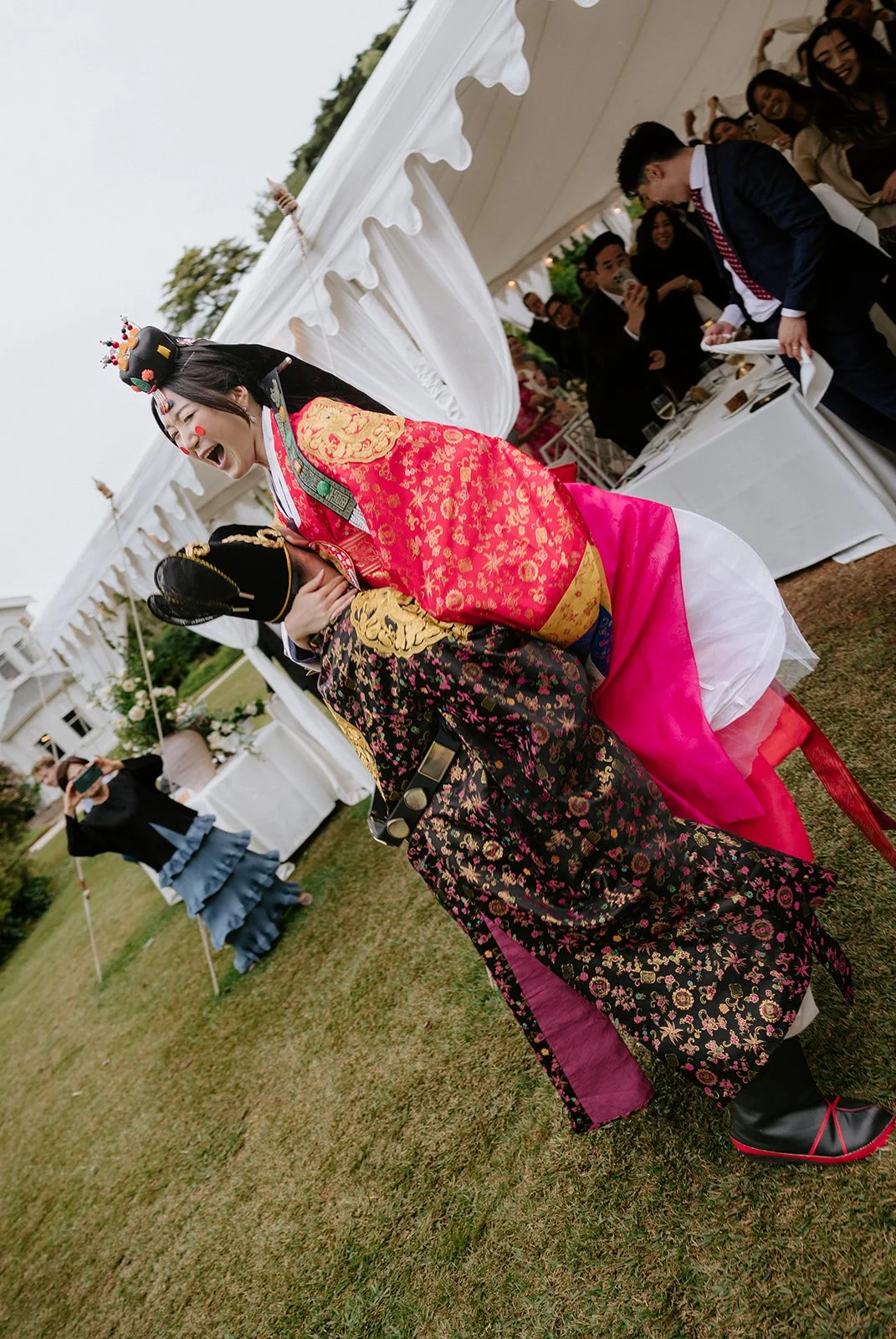 A woman dressed in traditional Korean wedding hanbok, laughing and leaning back while being supported by a man in a suit at an outdoor wedding reception with guests and white tents.