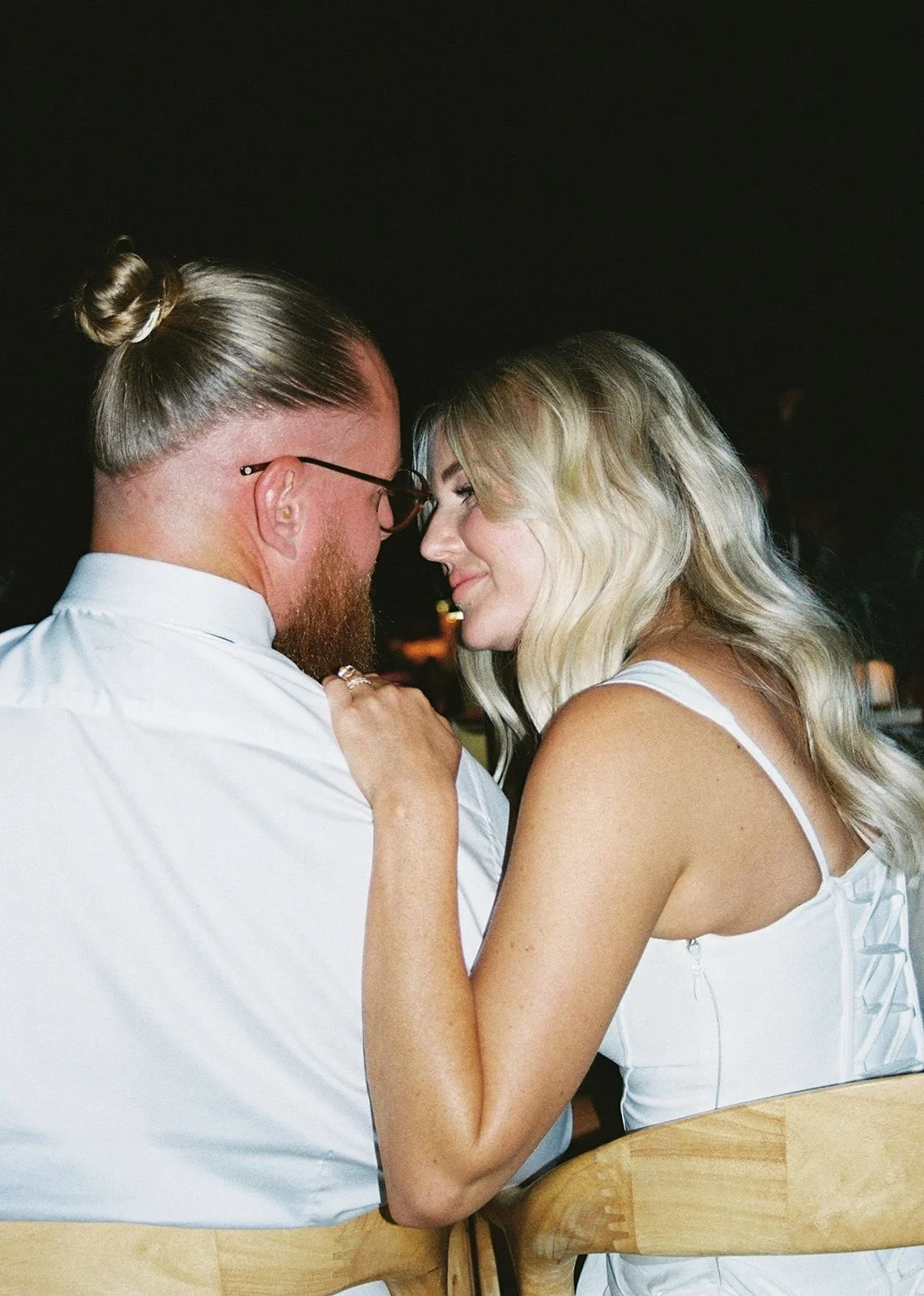 A couple faces close together at an indoor event, smiling softly at each other, with the woman’s hand on the man’s shoulder.