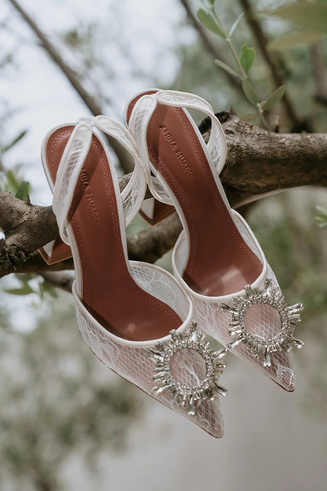 A pair of white lace high-heeled shoes with large rhinestone and silver buckle embellishments hanging from a tree branch.