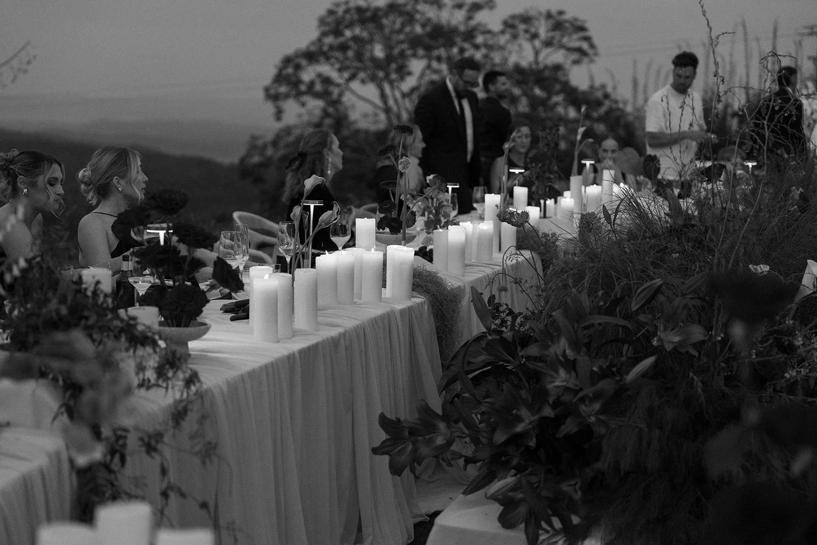 A black and white photo of a wedding reception with people seated at a long table decorated with candles and flowers, outdoors with trees in the background.