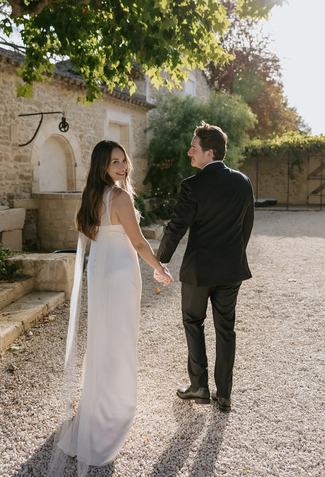 A couple dressed in wedding attire holding hands and walking outdoors on a gravel path, with a stone building and trees in the background, sunlight streaming through the leaves.