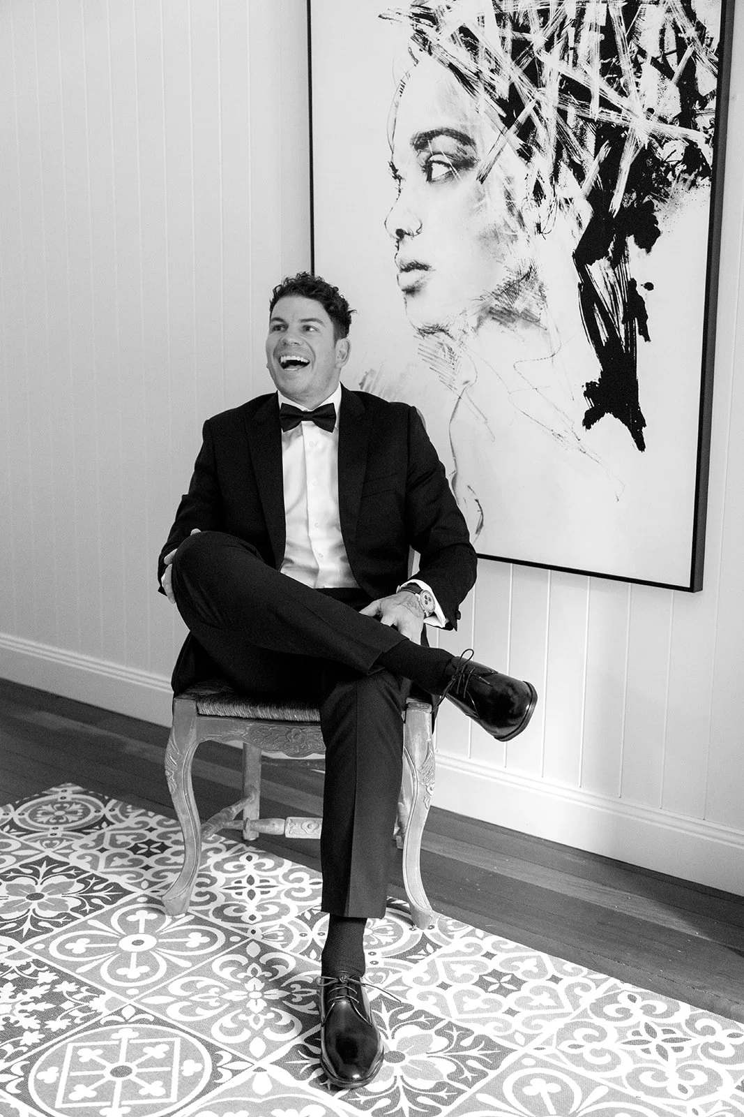 A man in a tuxedo sitting on a chair, smiling and laughing, in a room with a large abstract portrait painting behind him.