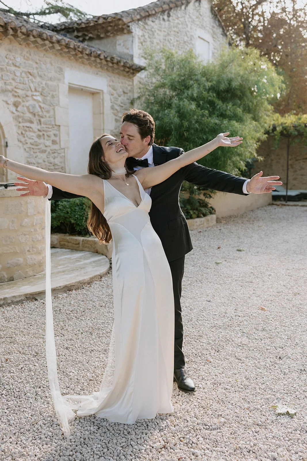 Bride and groom celebrating outdoors, smiling with arms outstretched, in front of a stone building with trees.