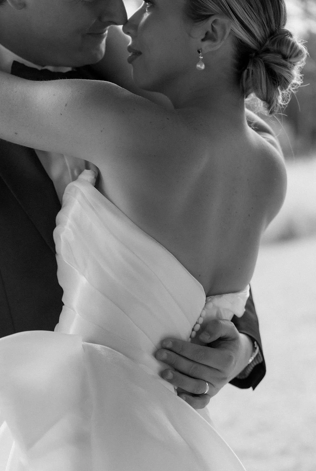 A black-and-white close-up of a bride and groom embracing, with the bride's face near the groom's nose, her hair in an elegant bun, wearing pearl earrings, and her wedding dress visible.