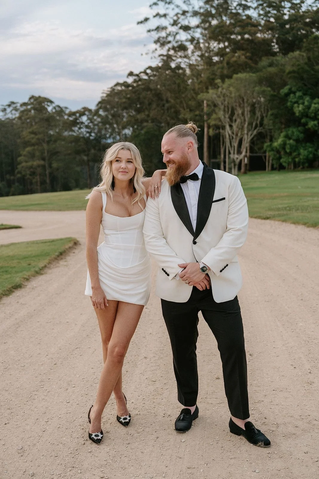 A man and woman in formal attire standing on a dirt path outdoors, with trees and grass in the background, smiling and looking at each other.