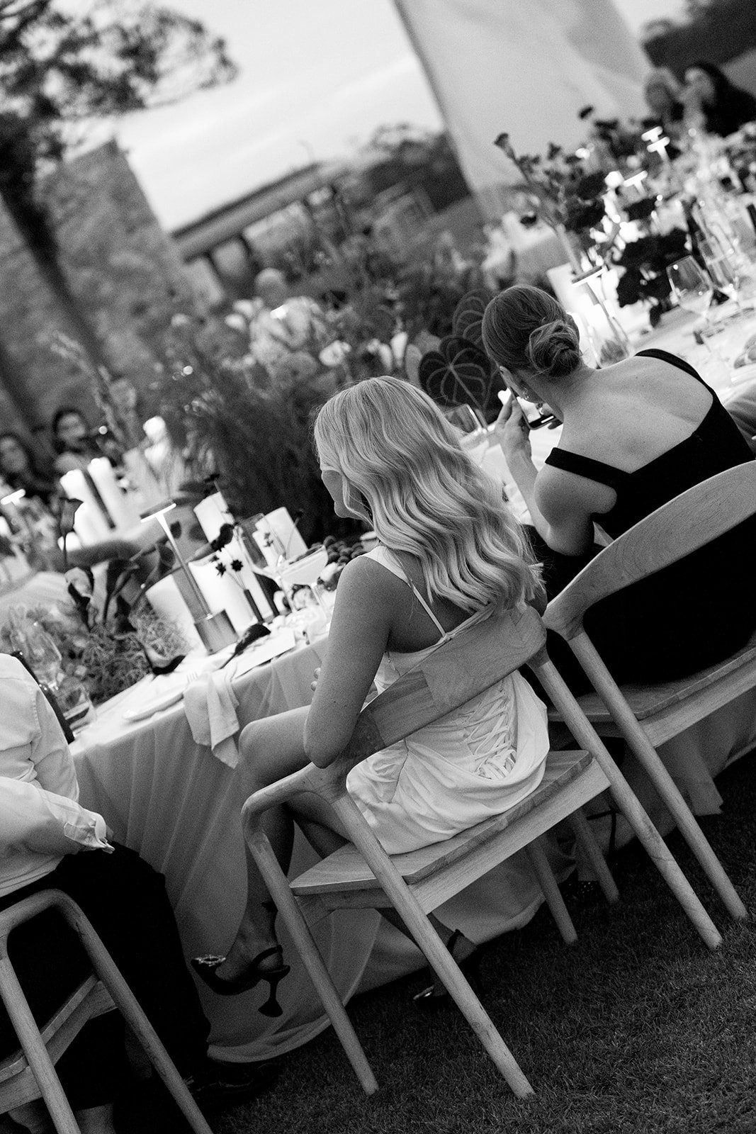 Two women sitting at a long outdoor banquet table decorated with flowers and candles, attending a festive event or celebration, with other guests and trees in the background.