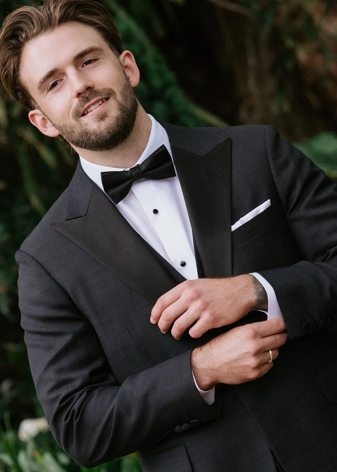 A man in a black tuxedo with a bow tie, white shirt, and pocket square, standing outdoors with greenery in the background.
