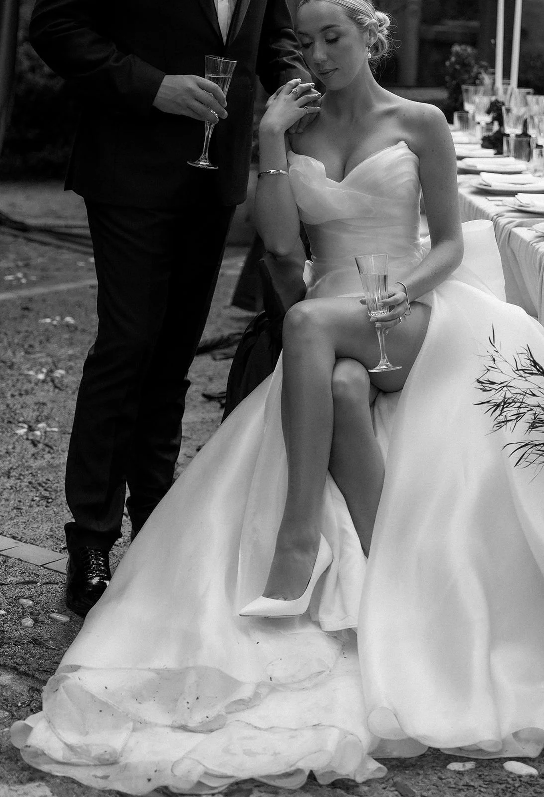 A bride in a wedding dress sitting with her legs crossed, holding a glass, while a man in a suit is standing next to her holding another glass. They appear to be at a wedding reception outdoors.