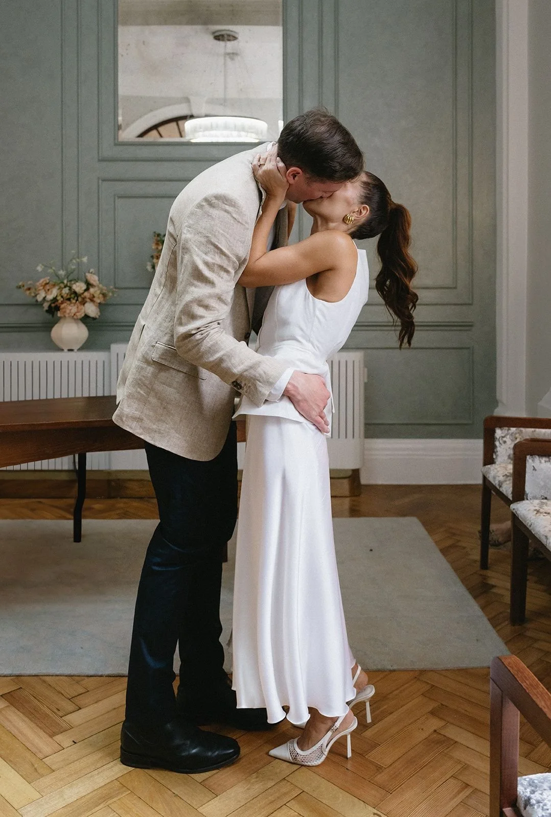 A man and woman are sharing a kiss in an indoor setting, with the woman on her heels, dressed in a white outfit, and the man in a beige blazer and dark pants. The woman is wearing high heels and gold earrings, and the background includes a wooden flo