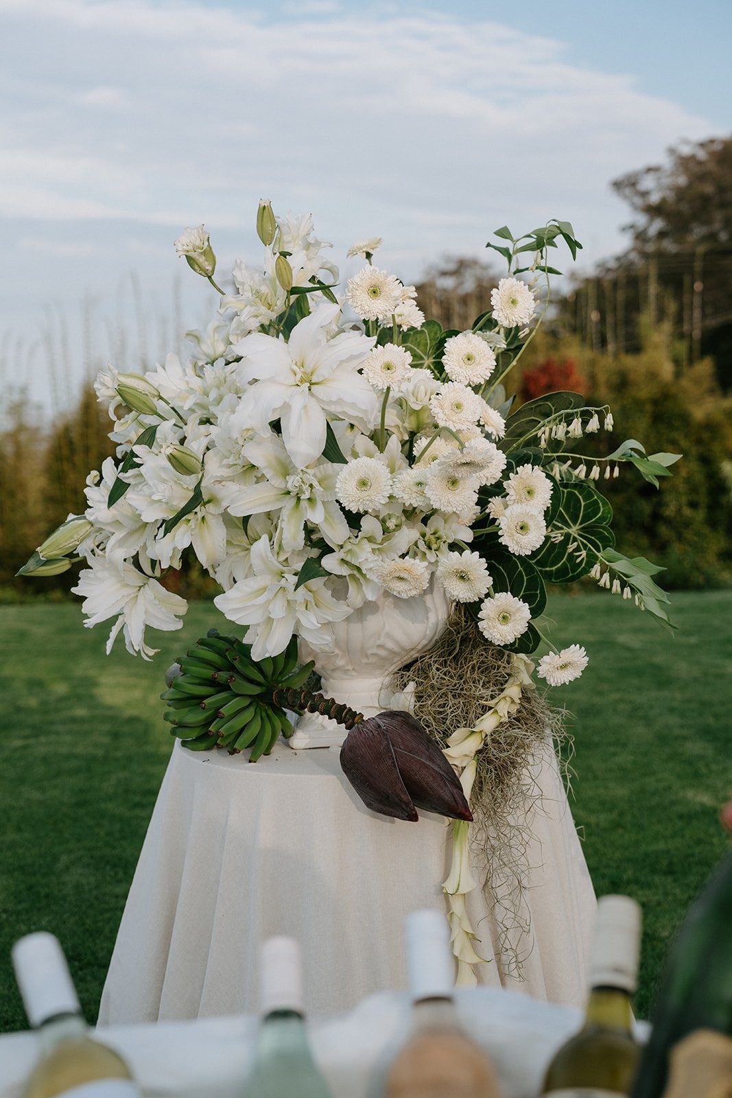 A floral arrangement of white flowers, including lilies, daisies, and other blooms, placed in a white vase on a table with a beige tablecloth, outdoors with greenery and trees in the background.