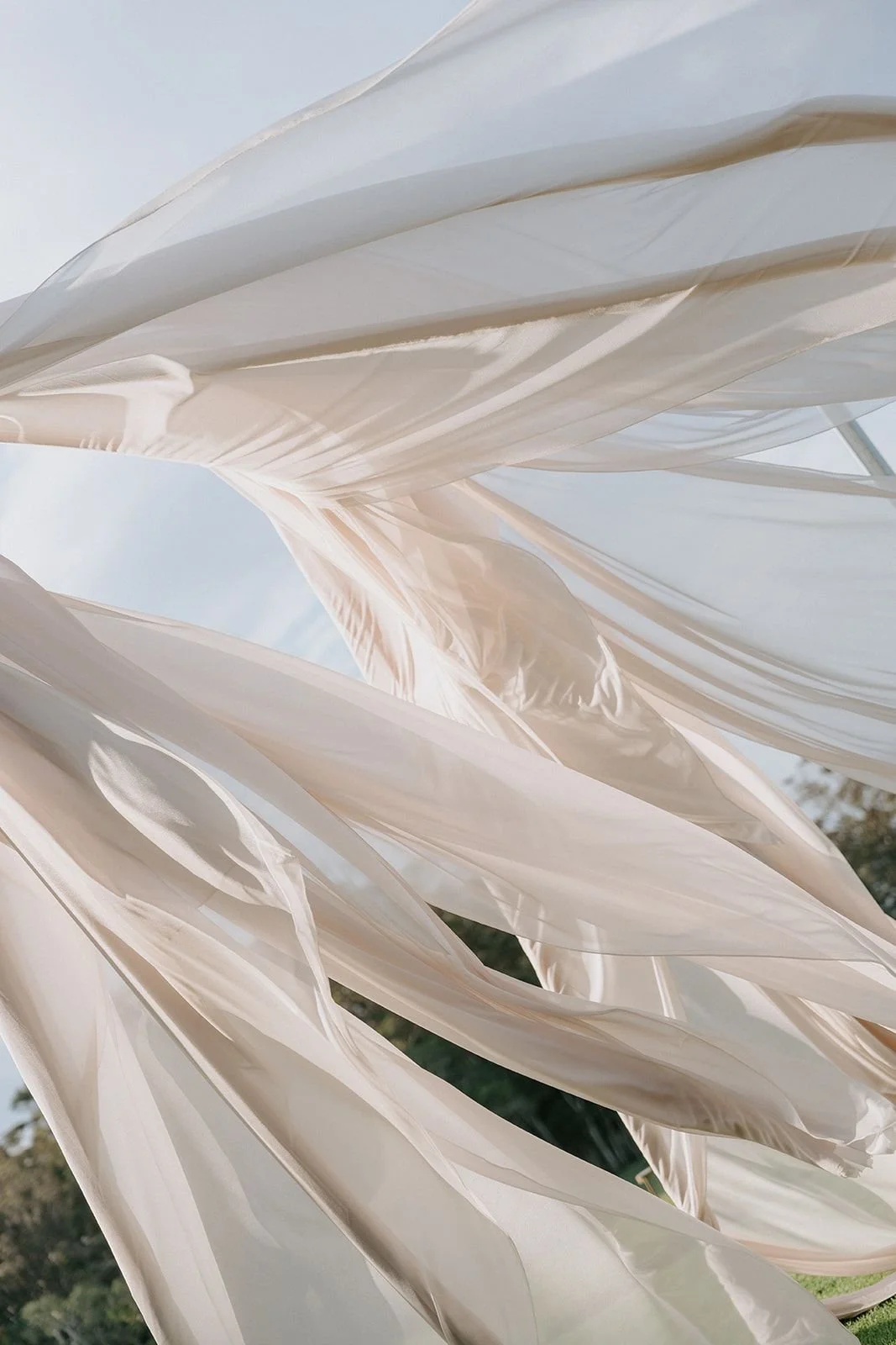 Close-up view of flowing, sheer white fabric outdoors with a blue sky and some greenery visible in the background