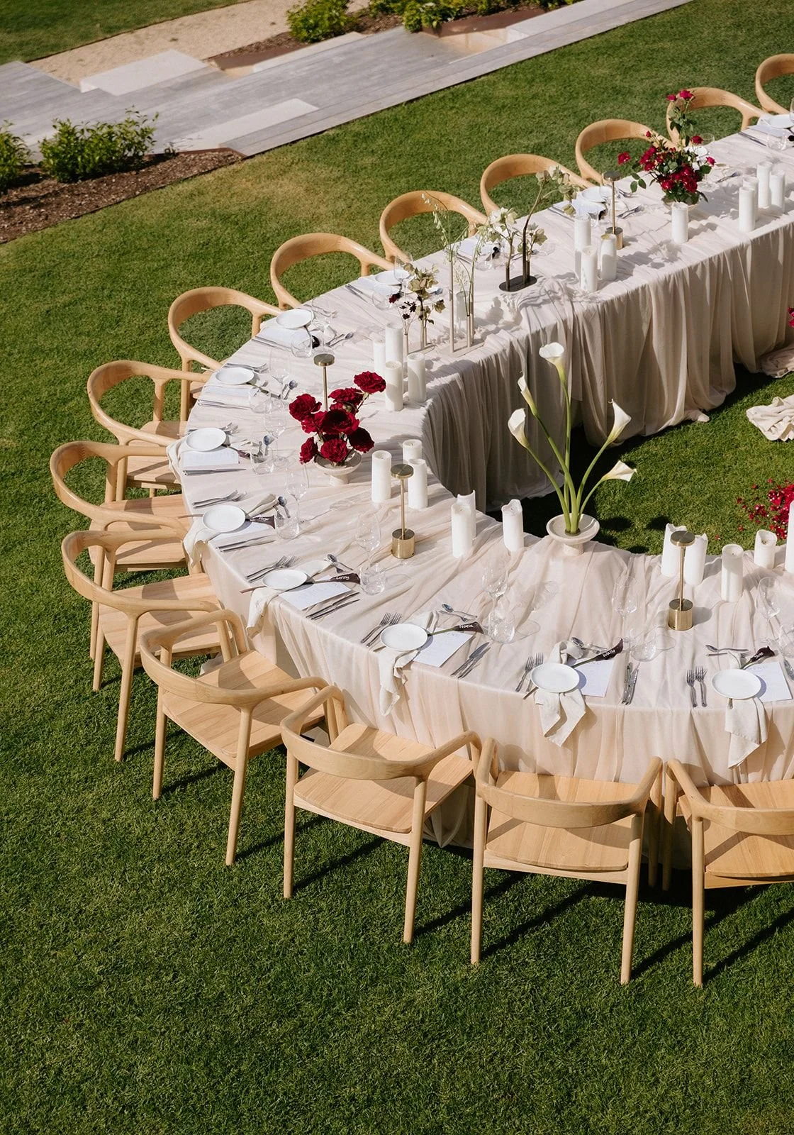 A long outdoor dining table set on a grassy area with beige chairs, white tablecloth, white plates, silverware, glassware, and floral centerpieces in white, red, and pink flowers.