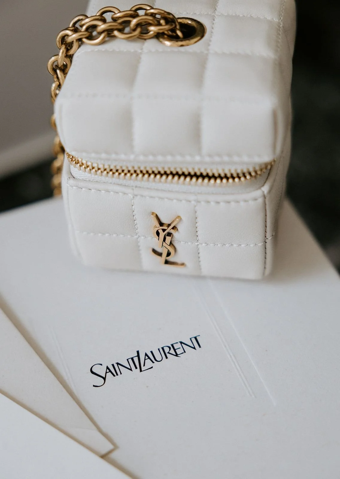 A white Saint Laurent mini handbag with gold accents and a chain strap, placed on top of white Saint Laurent shopping bags.