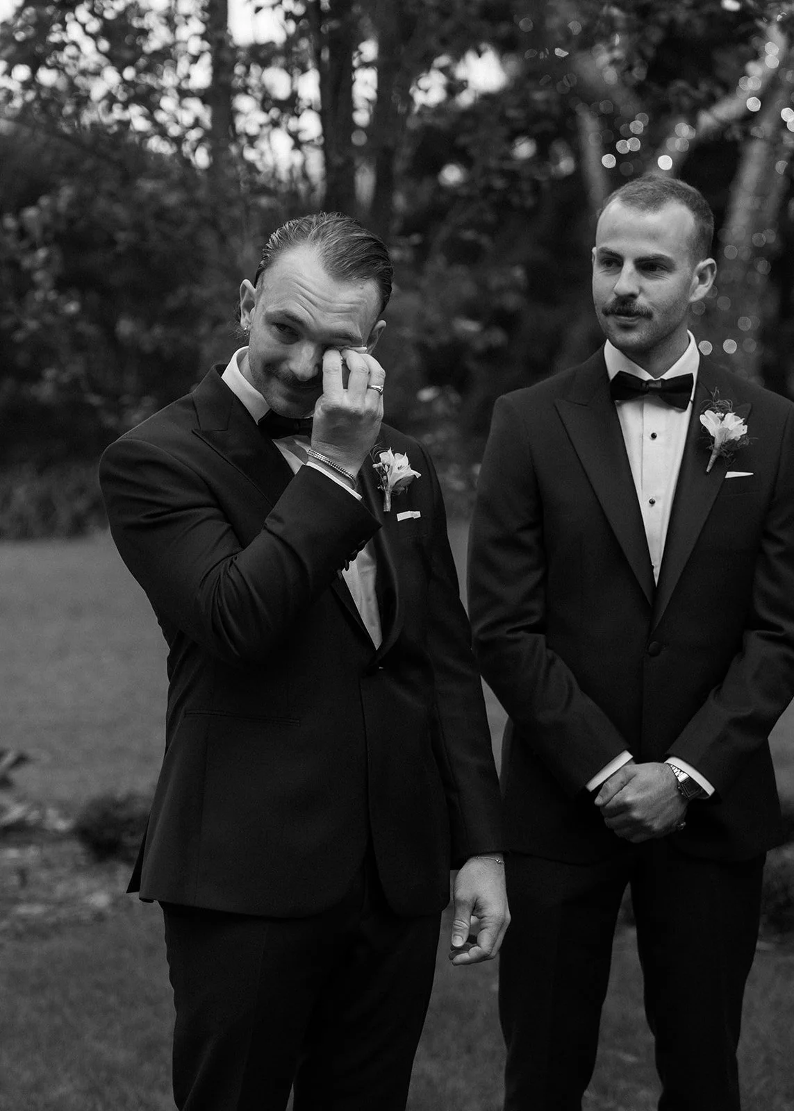 Two men in tuxedos outside, one wiping tears with a tissue, the other looking on. Both are wearing boutonnières, and the background has trees and faint string lights.