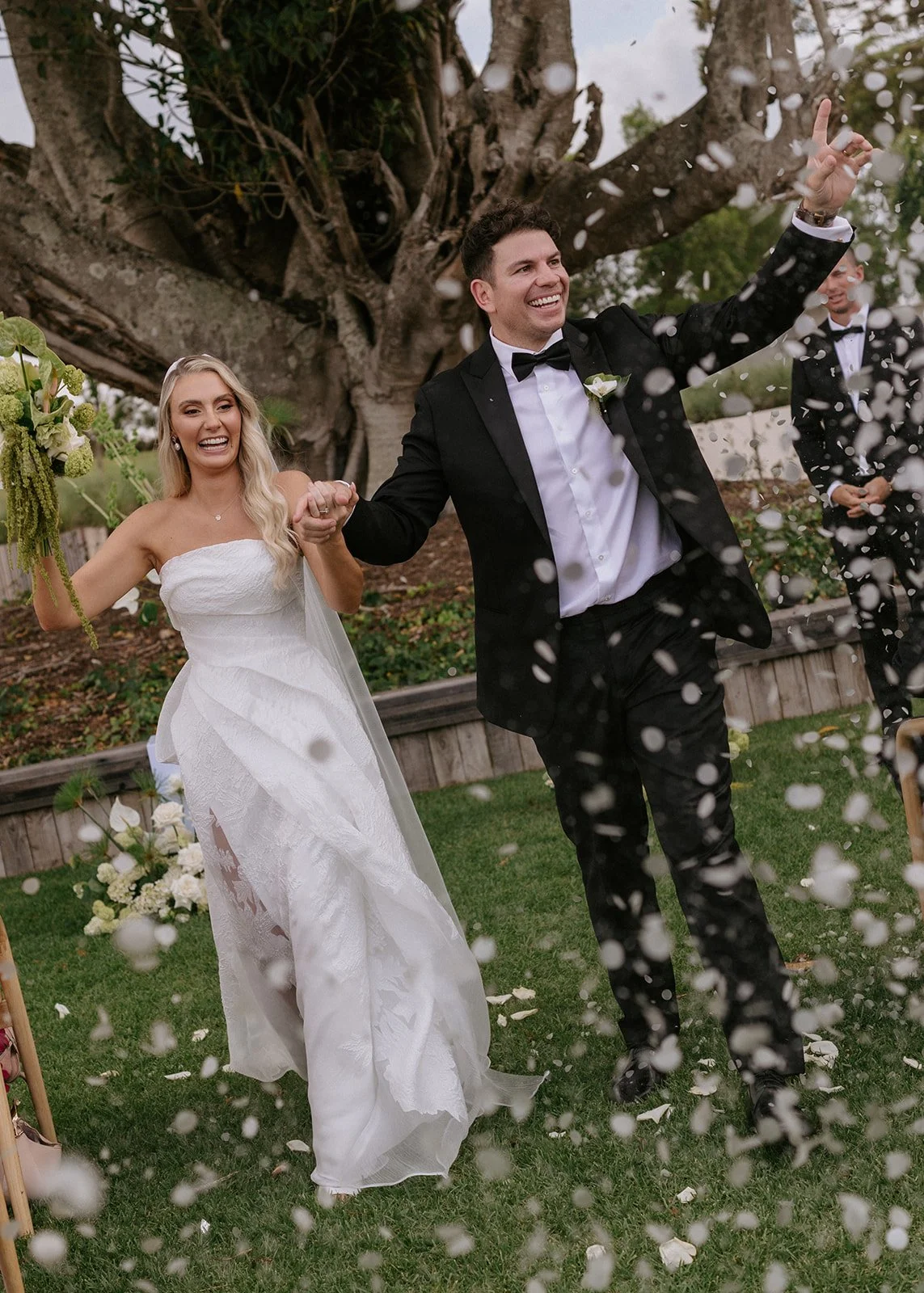 Bride and groom celebrating their wedding outdoors under a large tree, with white confetti falling around them, the bride in a strapless white gown and the groom in a black tuxedo, holding hands and smiling.