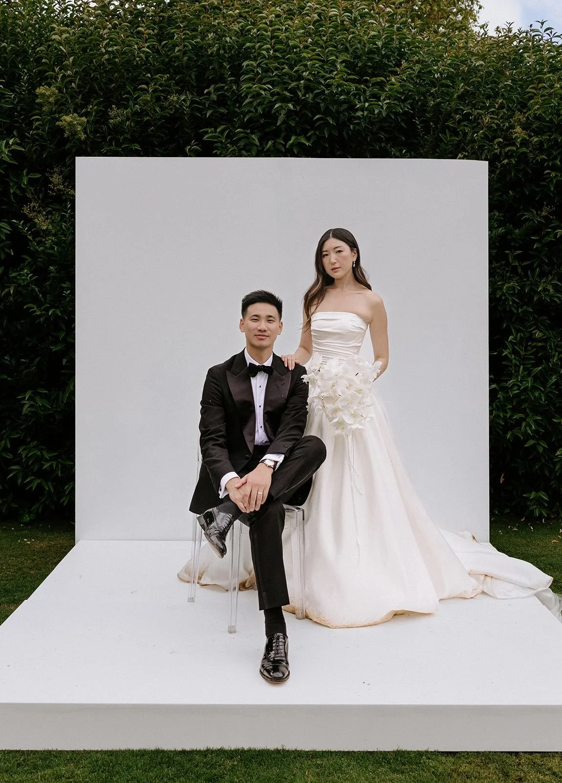 A couple dressed in wedding attire posing outdoors in front of a white backdrop with greenery. The groom is seated on a transparent chair, wearing a black tuxedo, and the bride is standing beside him in a strapless white wedding gown, holding a bouqu