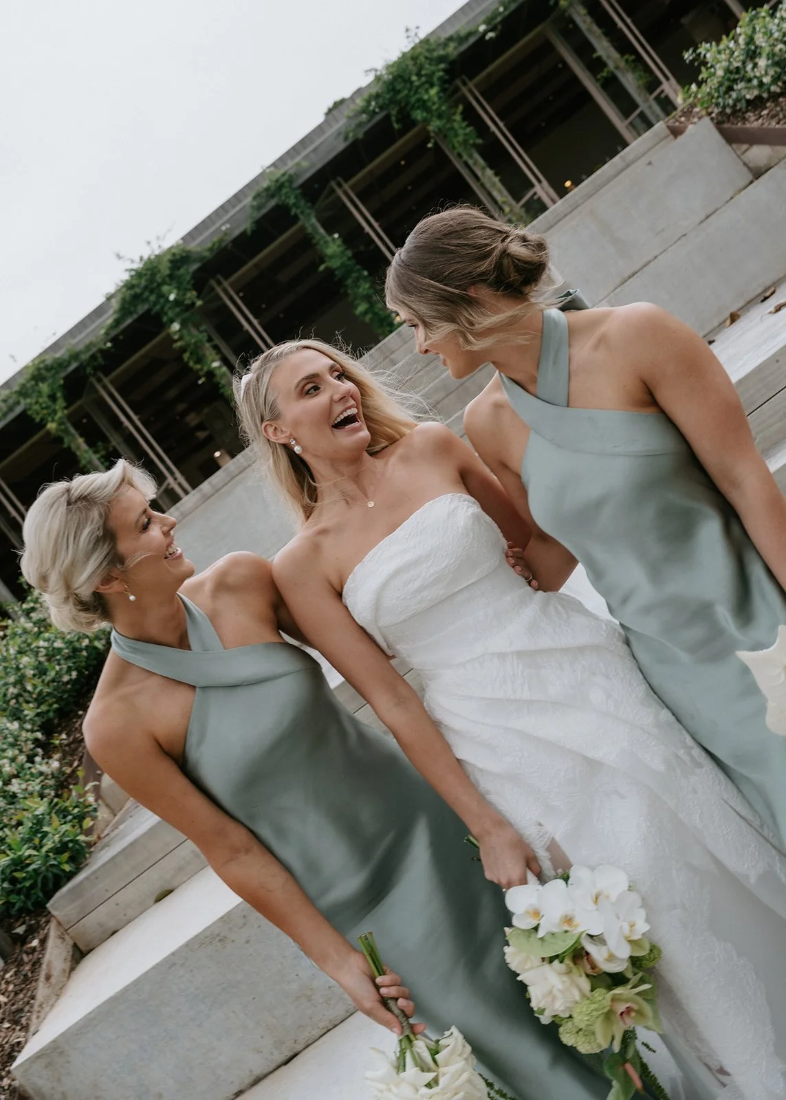 A bride in a white wedding dress standing with two bridesmaids in green dresses, all smiling and enjoying a moment together outdoors, holding bouquets of white flowers.