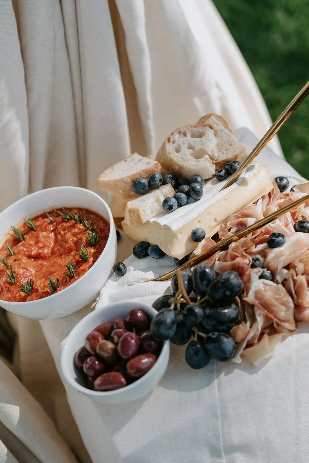 A cheese and charcuterie board with bread, grapes, cheese, and a bowl of tomato-based dip, set on a white tablecloth outdoors.