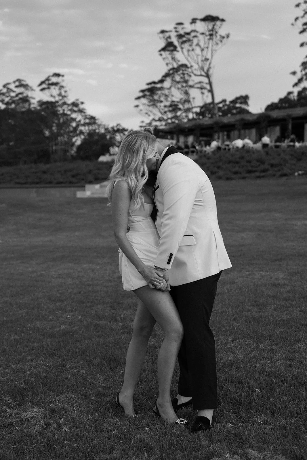 A couple kissing outdoors, with the woman wearing a dress and the man in a light-colored suit, standing on grass with trees and a building in the background.