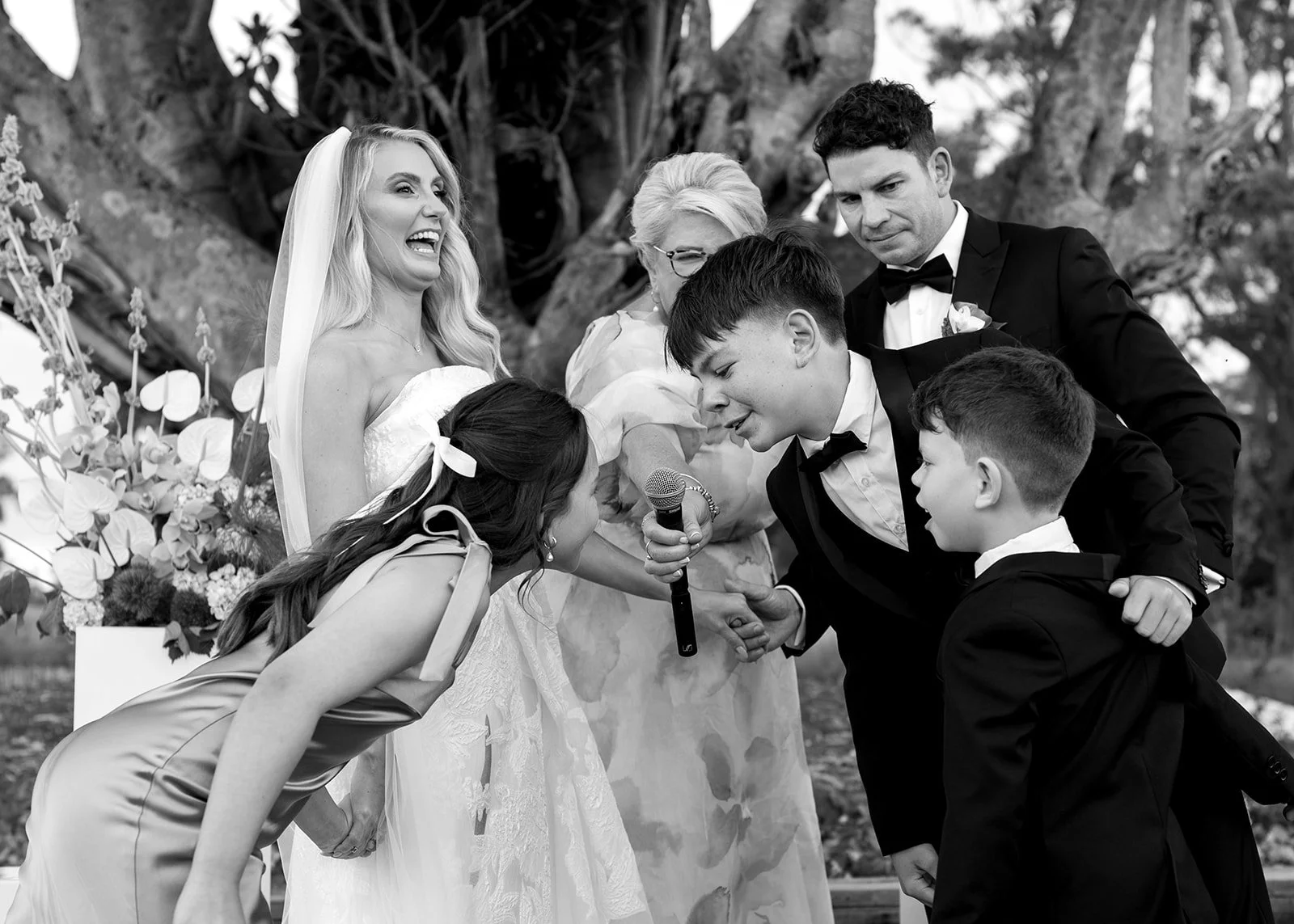 Black and white photo of a wedding scene with seven people. Two women in wedding dresses, one laughing and holding a bouquet, the other leaning forward. Four men in tuxedos, one holding a microphone, two with hands clasped, all engaged in a moment of