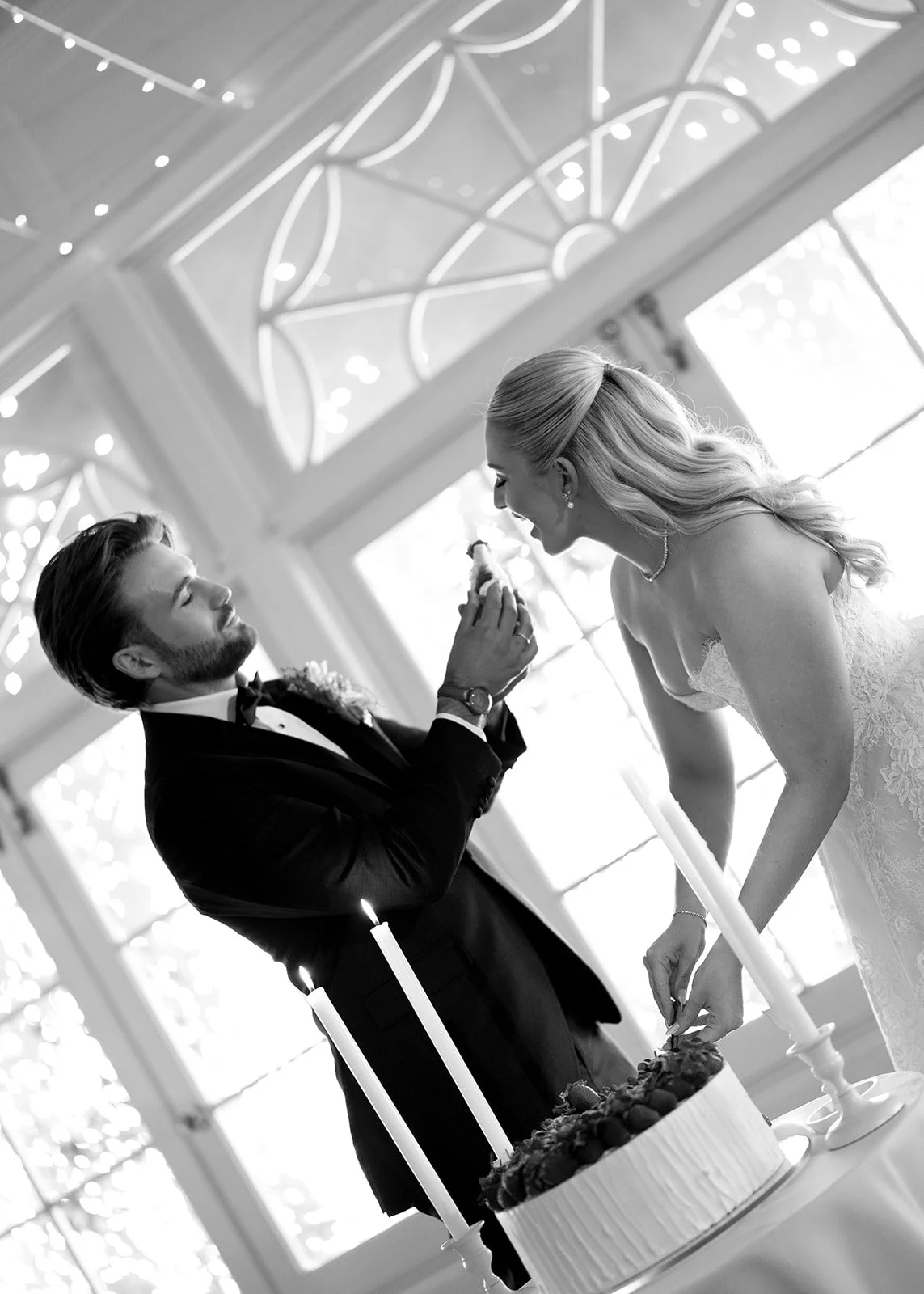 A bride and groom cutting their wedding cake during a celebration, with candles and decorative windows in the background.
