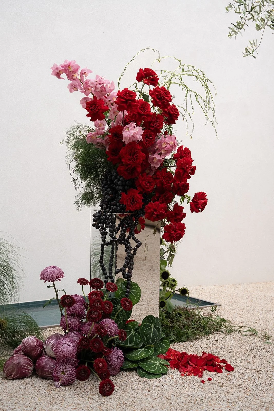 An arrangement of various flowers including pink and red blooms, black grapes, and green leaves, set against a plain background.